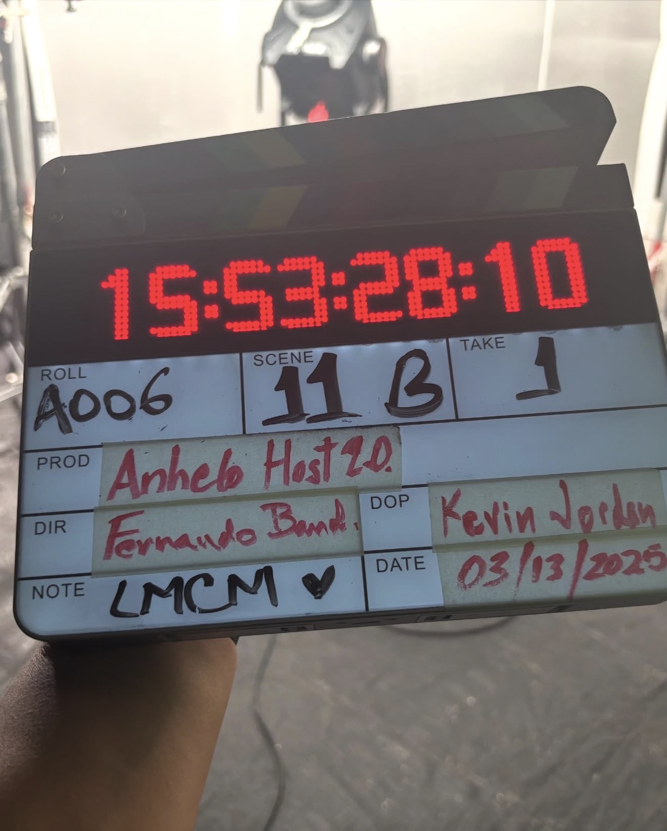 Clapperboard with red digital clock showing 15:53:28:10, handwritten notes, and a blurred background of a hallway.