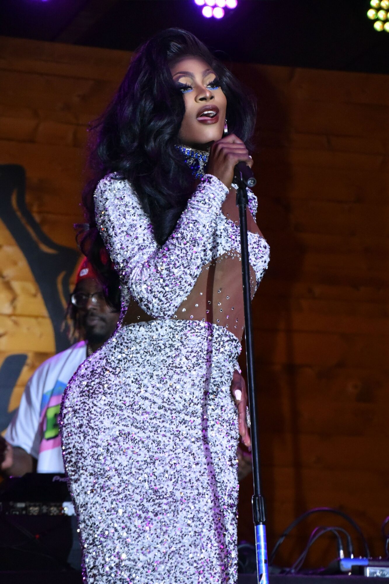 Woman with dark hair singing into a microphone on stage, wearing a sparkly dress, with a wooden background.