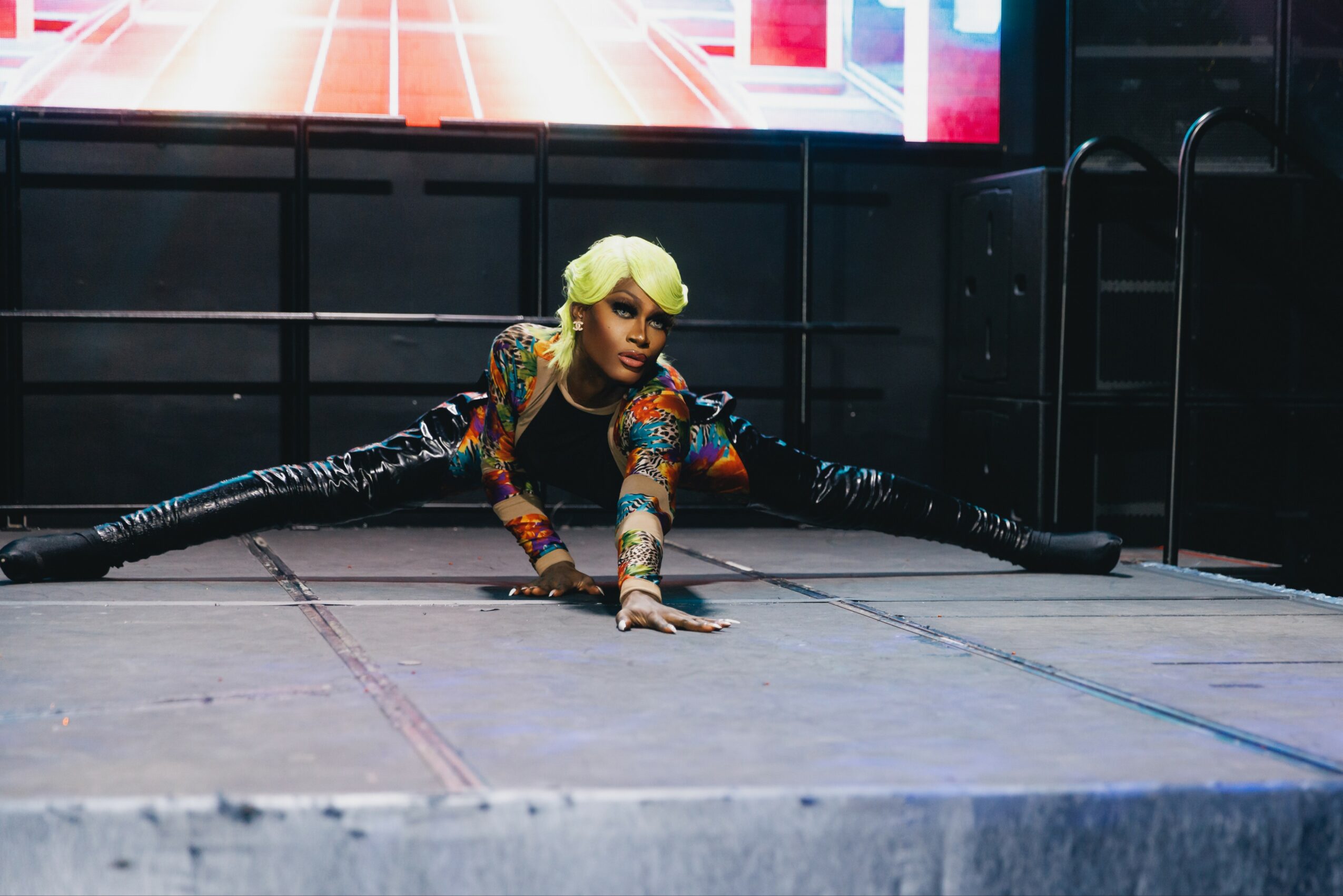 Person with blonde hair in a colorful outfit performing a split on stage, with arms extended forward and a digital screen in background.