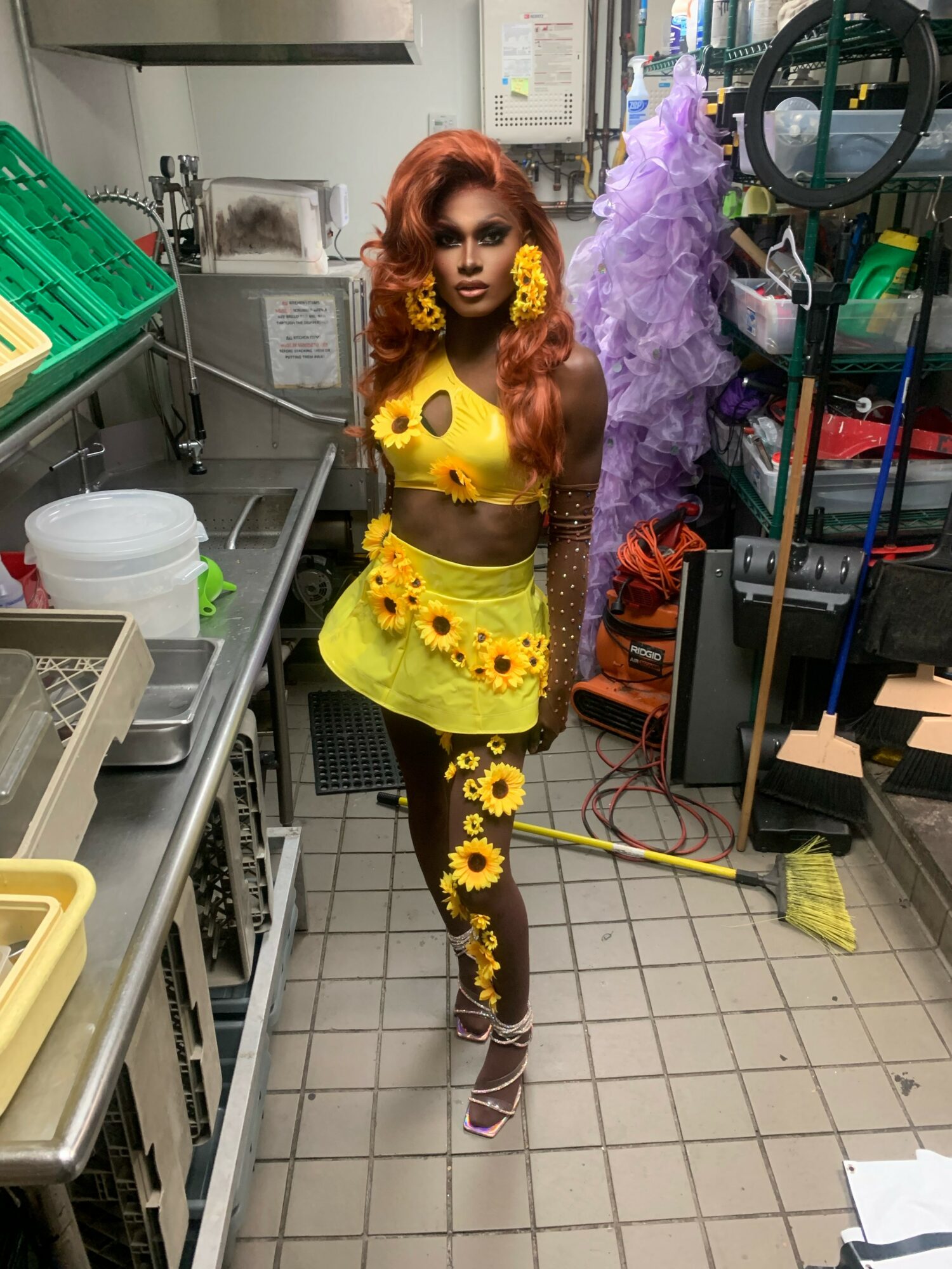 Person with long red hair in a yellow floral outfit standing in a kitchen or storage room.