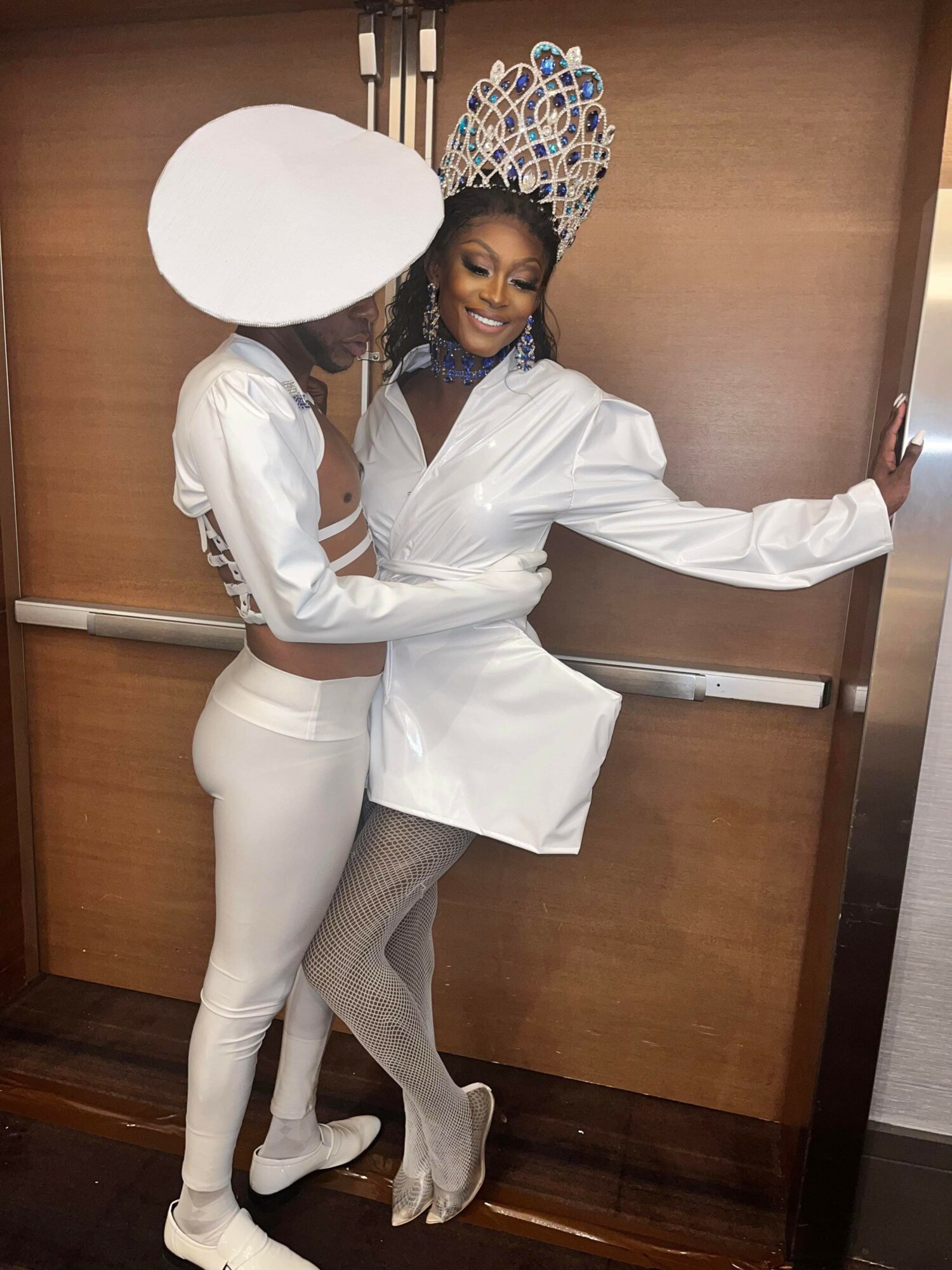 Two women in stylish outfits pose inside an elevator, one wearing a crown and the other with a large white hat.