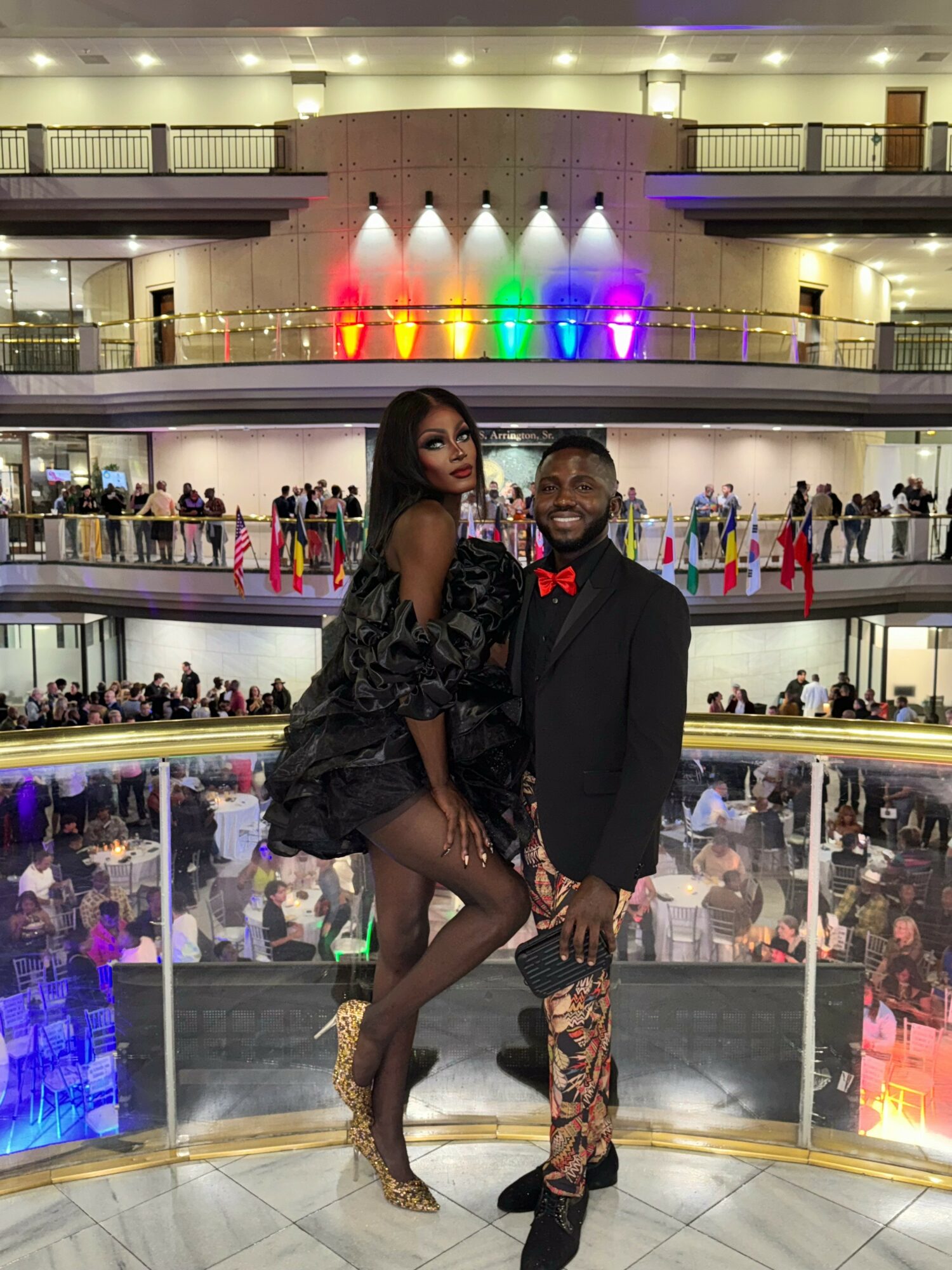 A man and woman pose together in a multi-level shopping mall with colorful lights and crowds in the background.