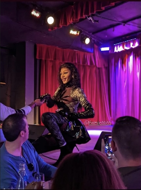 Performer on stage with red curtains, smiling, wearing a shiny outfit, audience watching, purple stage lighting, holding microphone.