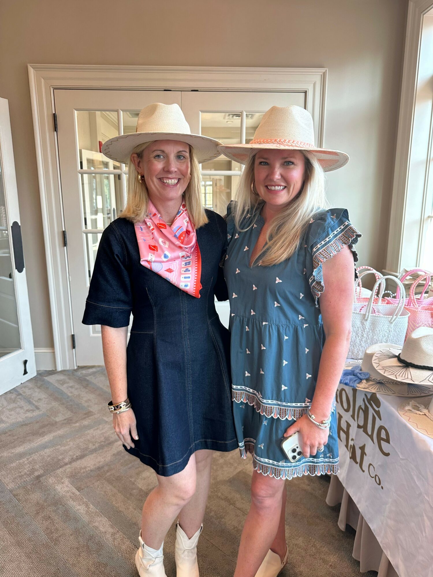 Two women in cowboy hats and casual outfits standing indoors, smiling, with a table and window in the background.