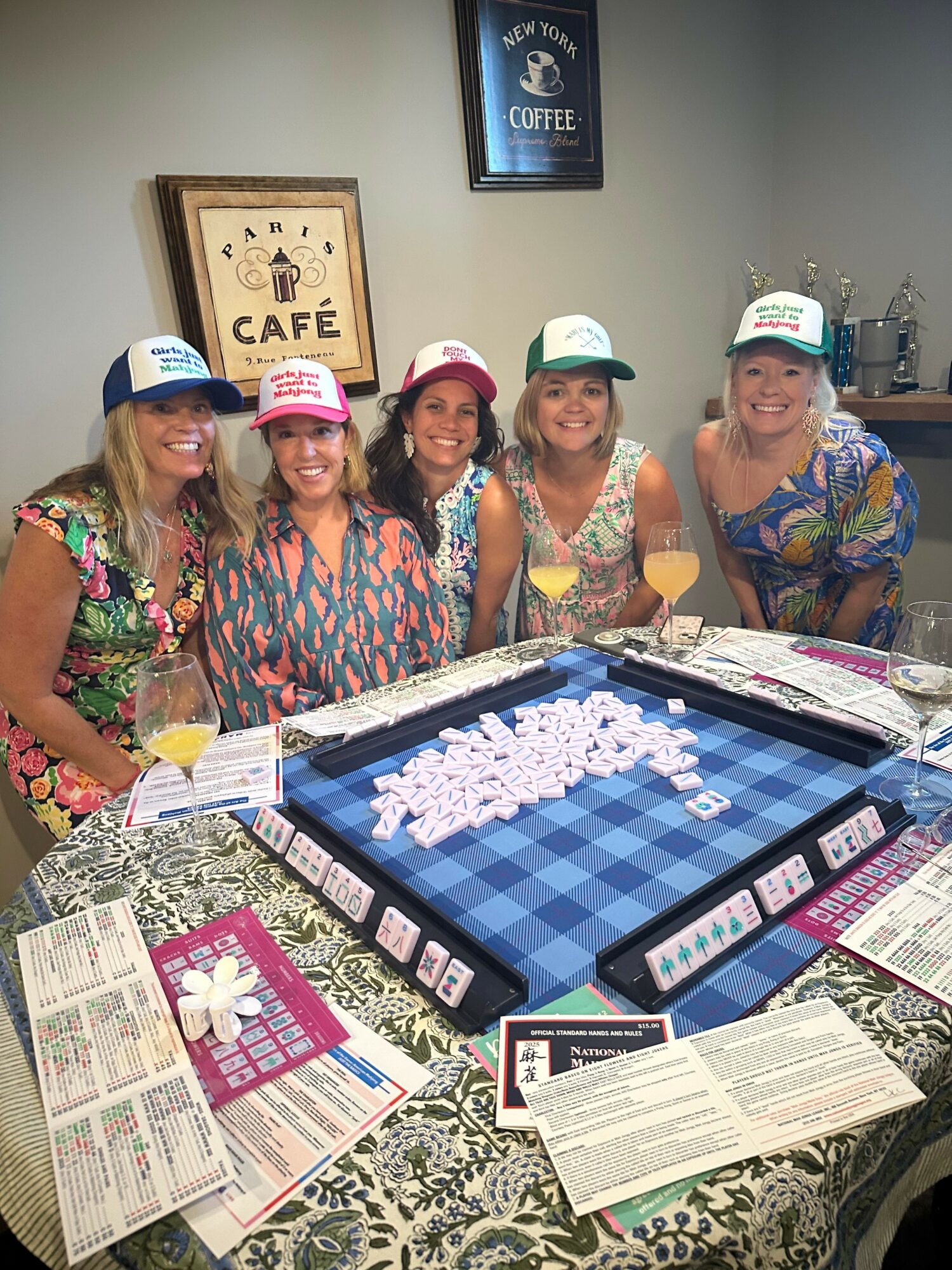 Group of five people wearing colorful shirts and hats playing a board game at a table with papers and cards.