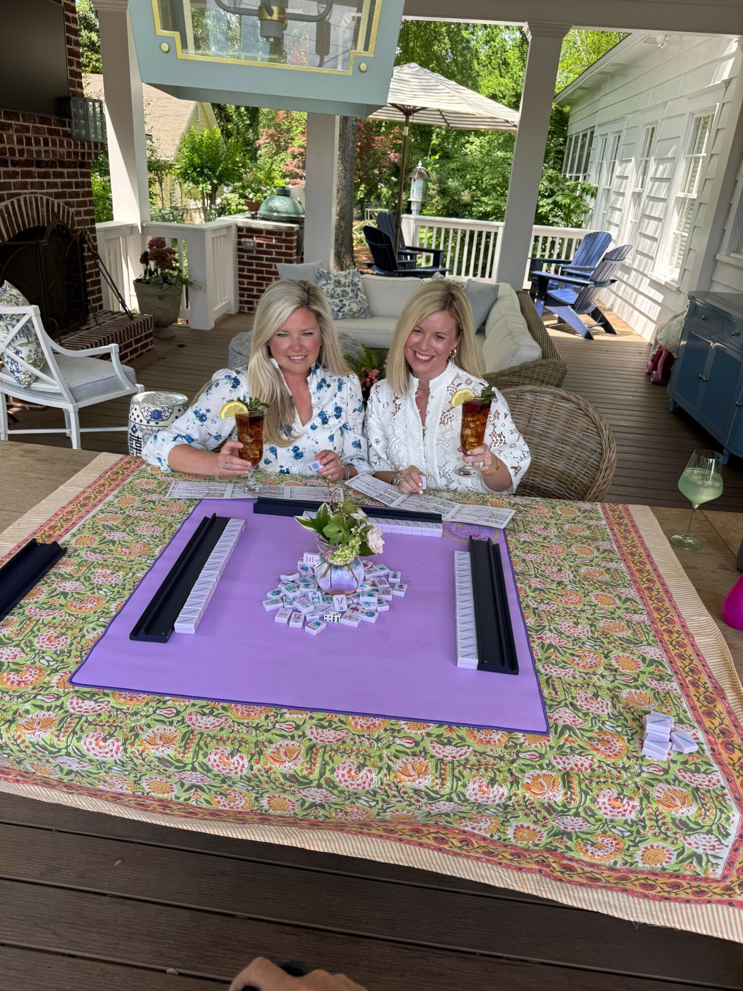 Two women sit at a table with a purple mat, flowers, and small objects, in a bright room with large windows and outdoor view.