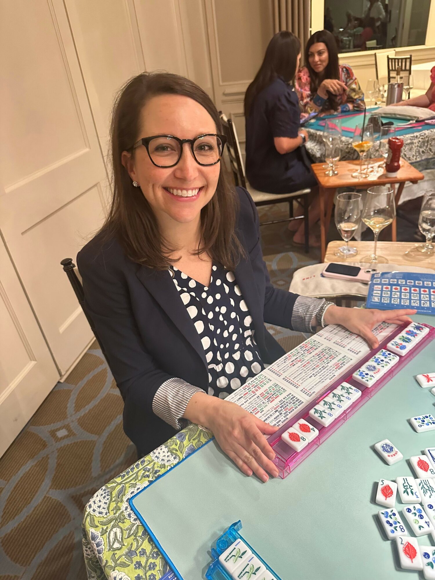Woman with glasses smiling at table with Mahjong tiles and score sheet, in a room with other people and tables.