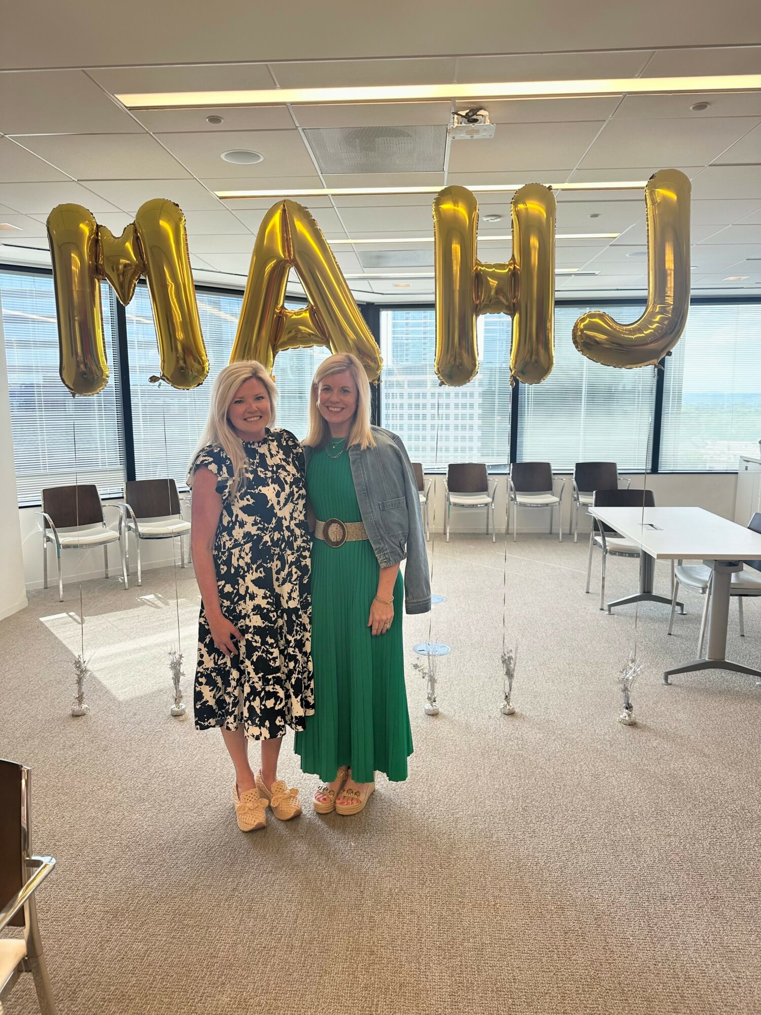 Two women standing in a room with large gold balloons spelling 'MAHJ' behind them, windows with a view outside, chairs and tables.
