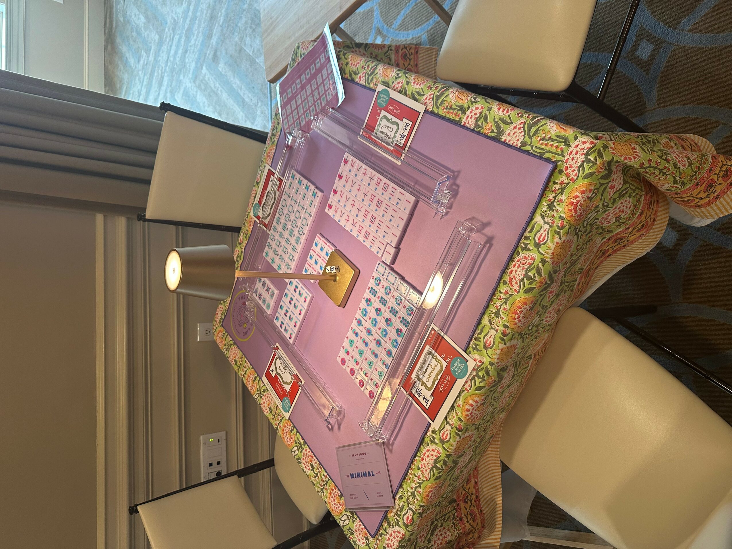 Table with a pink game board, cards, and game pieces, surrounded by chairs in a room with a window and curtains.