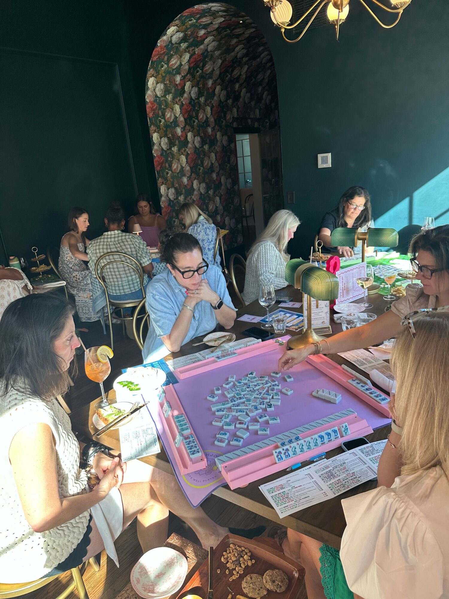People gathered around a table playing a board game, with drinks and snacks, in a room with dark green walls.