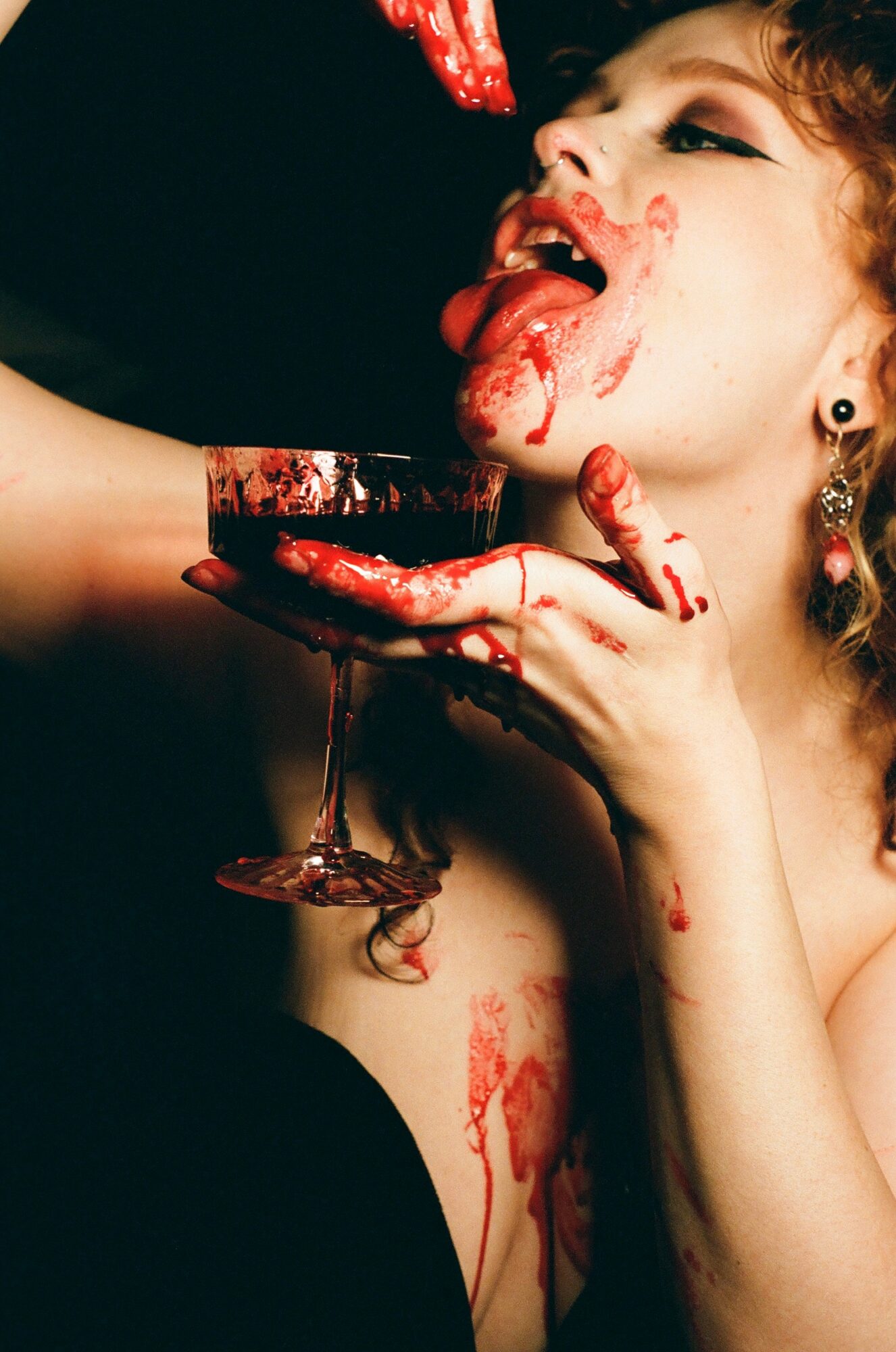 Person with blood on face and hand, holding a glass with blood, licking lips, with blood on arm and face.
