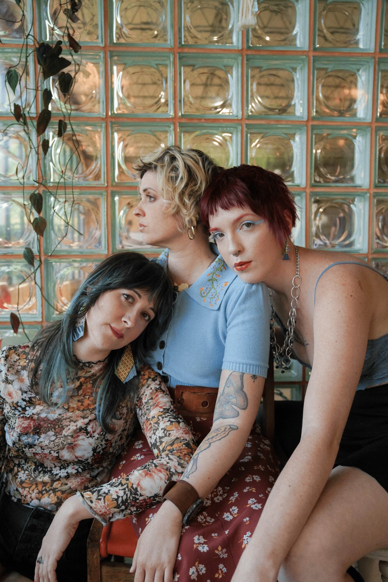 Three women sit together indoors near a glass block wall, with one woman leaning on another, all looking at the camera.