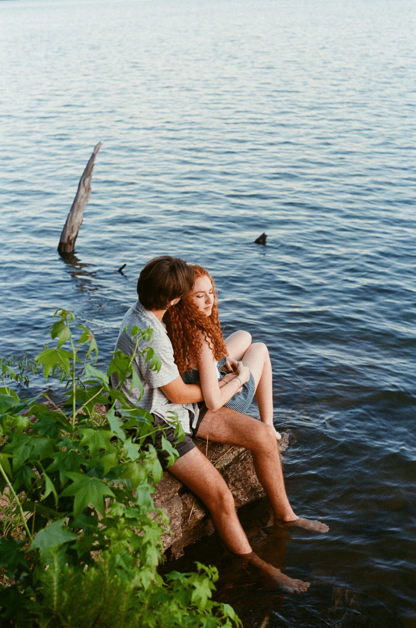 Two children sit on a rock by the water, with greenery nearby and a tree branch in the water.