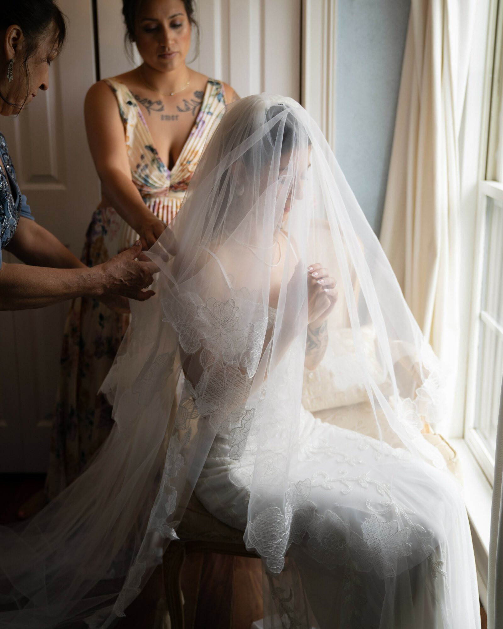 Bride sitting by window with veil, two women assisting her, sunlight streaming in, indoor setting.