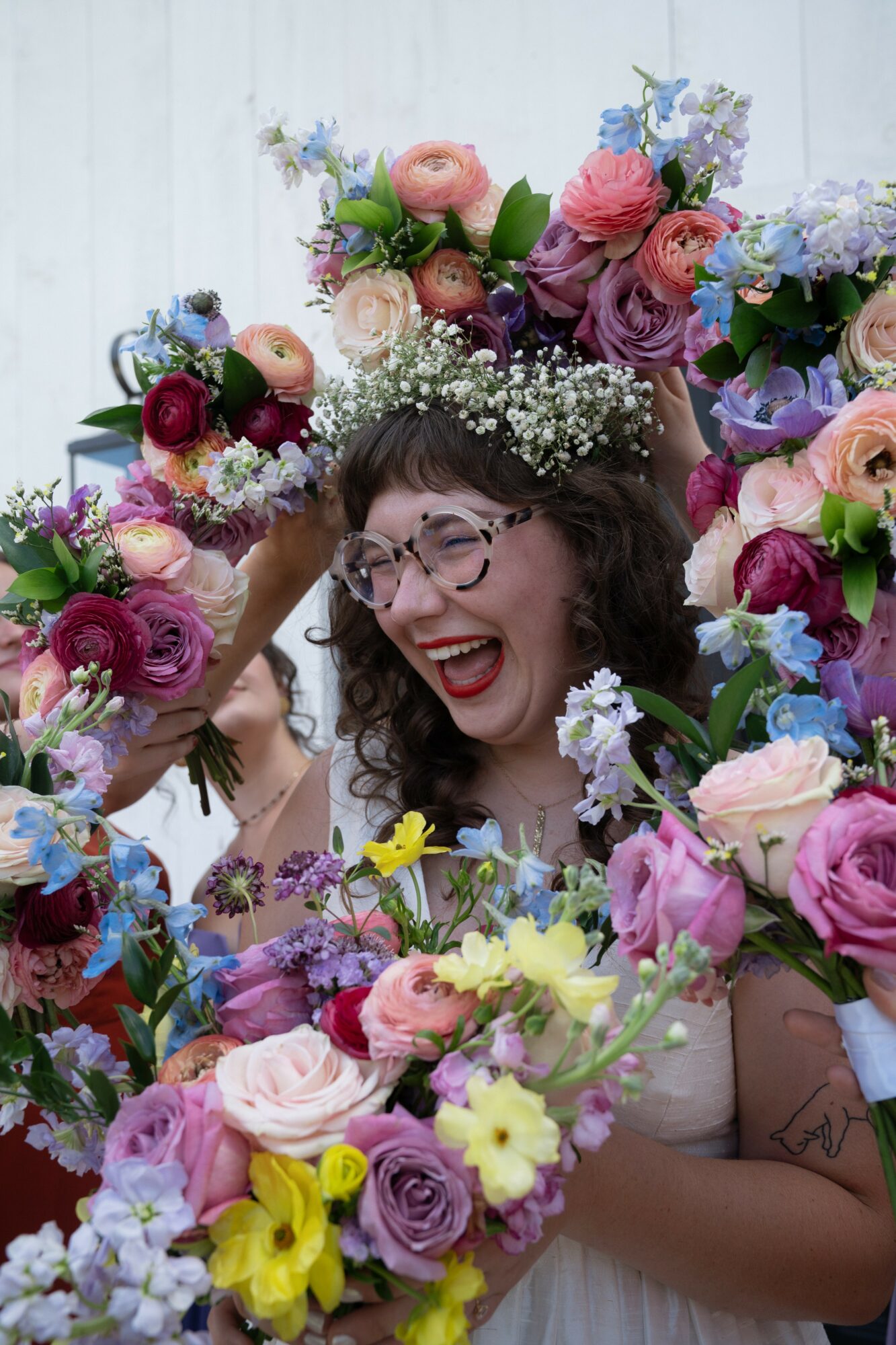 Woman with glasses smiling surrounded by colorful flowers and greenery.