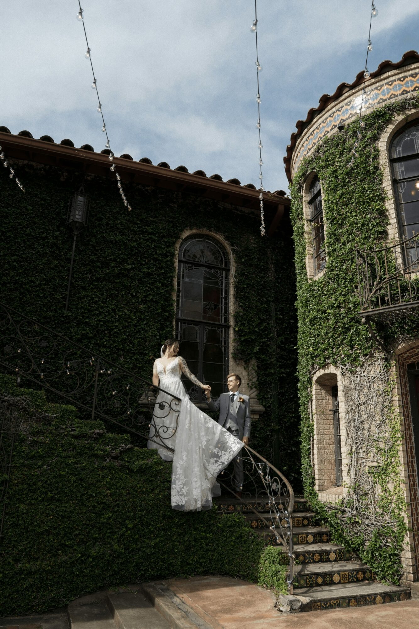 Bride and groom standing on stairs outside a building covered in ivy, with string lights overhead.