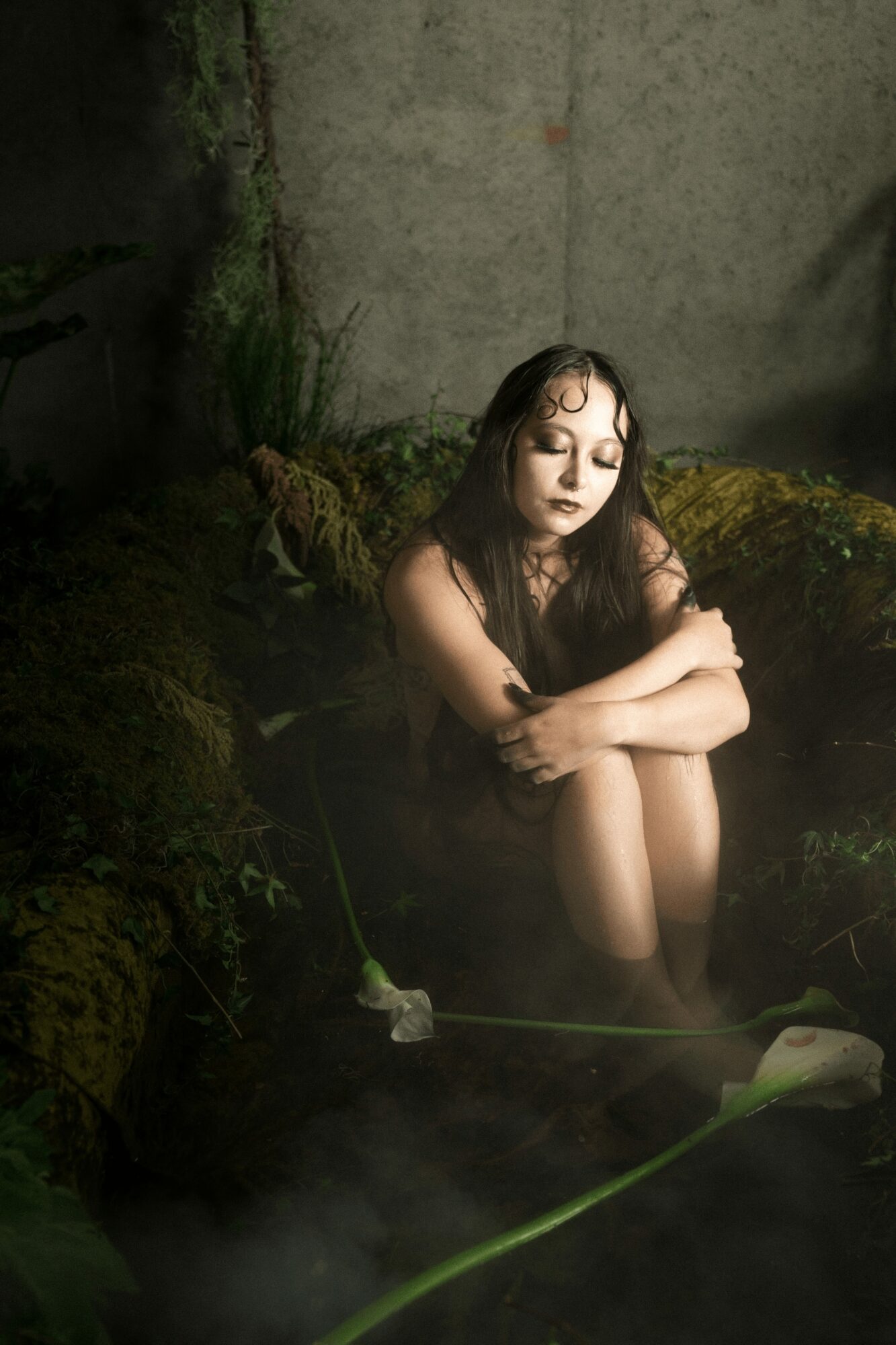 Young woman sitting with arms crossed, eyes closed, surrounded by moss and rocks in a natural setting.