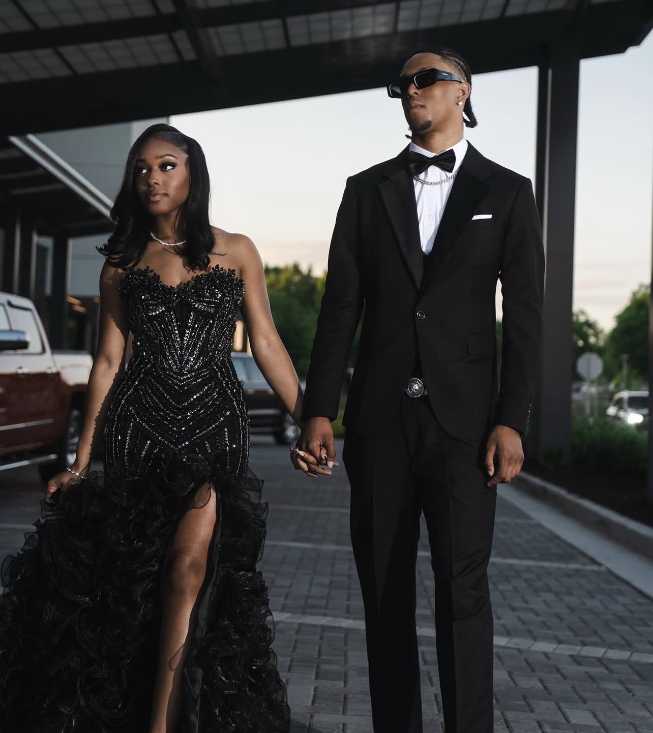 A man and woman in formal attire holding hands outdoors, woman in a black gown and man in a tuxedo, under a modern structure.