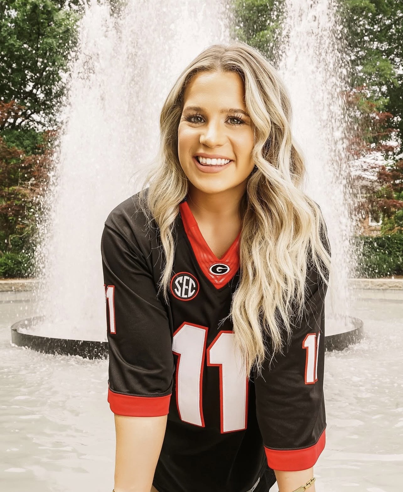 Young woman with long wavy blonde hair smiling in front of a fountain, wearing a black sports jersey with red and white details.
