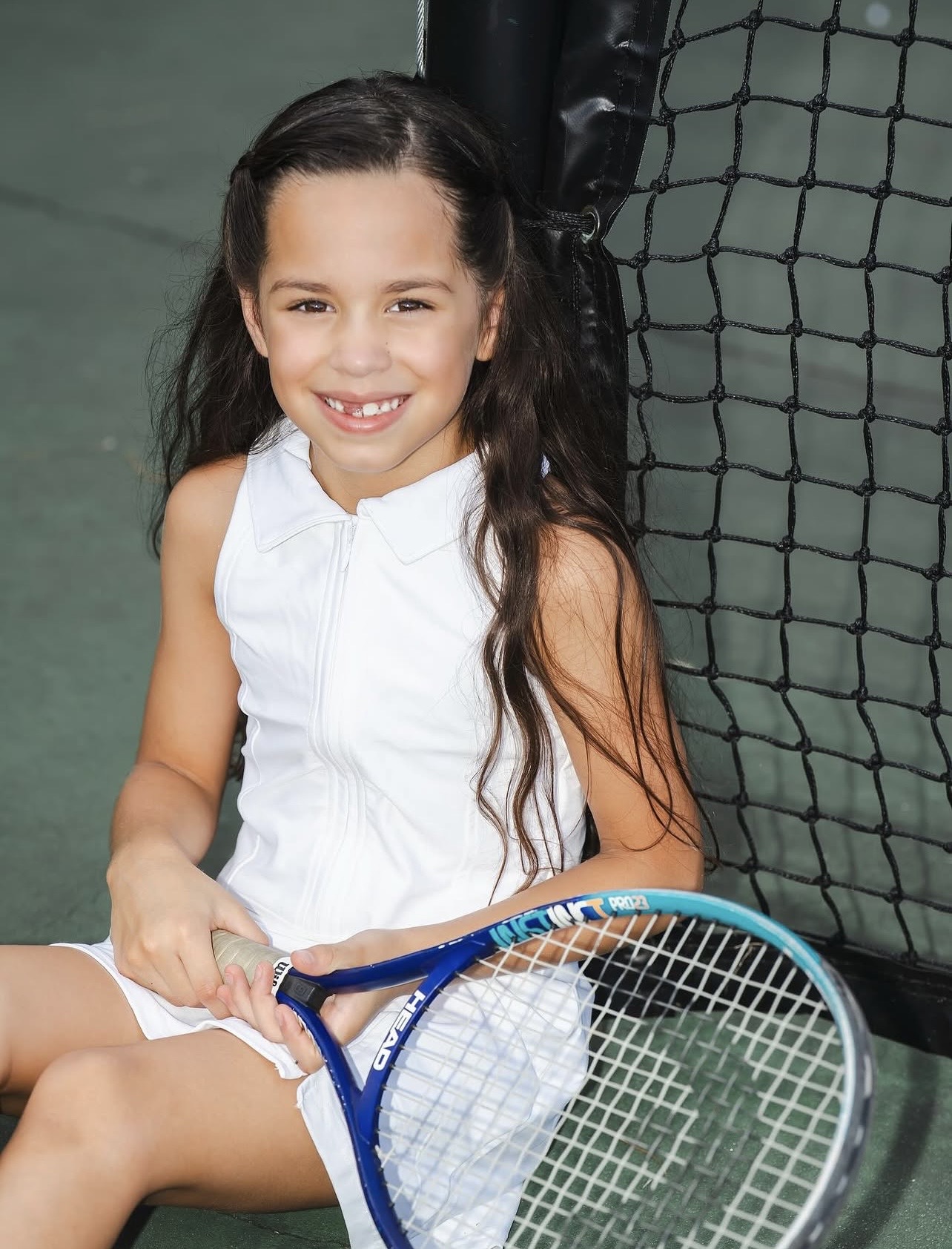 Young girl with long dark hair smiling, sitting on a tennis court holding a tennis racket, wearing a white sleeveless top.