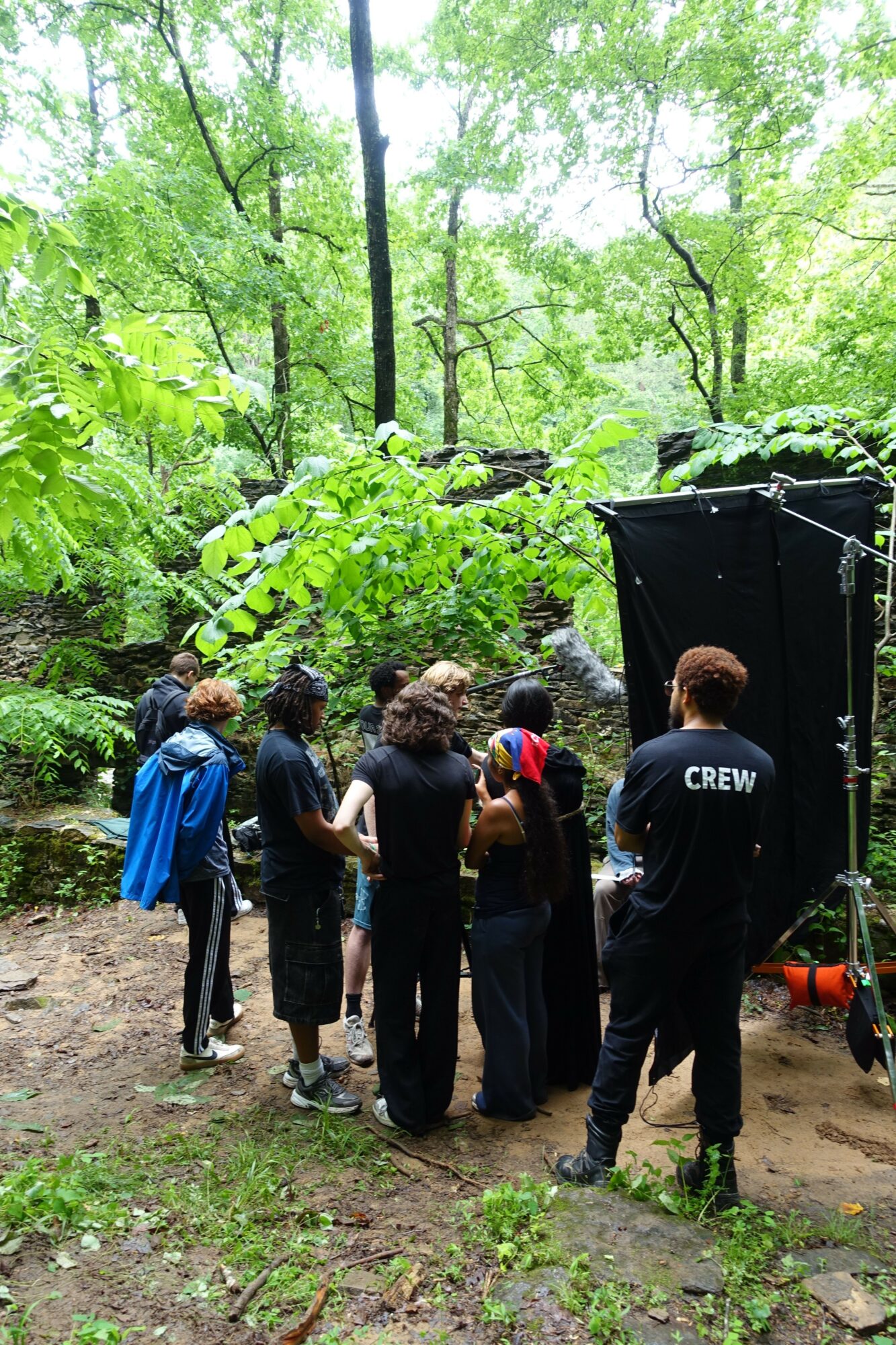 Group of people standing outdoors in a green forest, some wearing headphones, near a black tent or structure, with trees and foliage around.