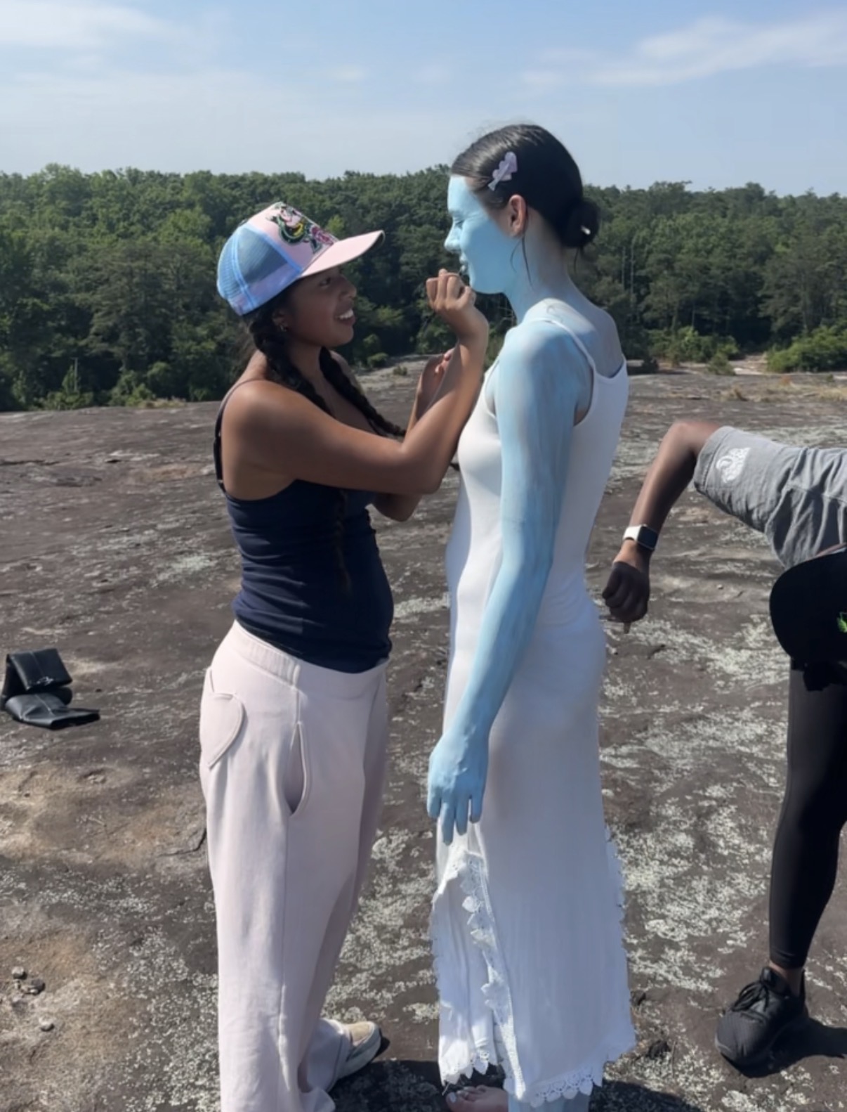 Two women stand outdoors, one applying face paint to the other, who is dressed in a white costume with blue body paint.