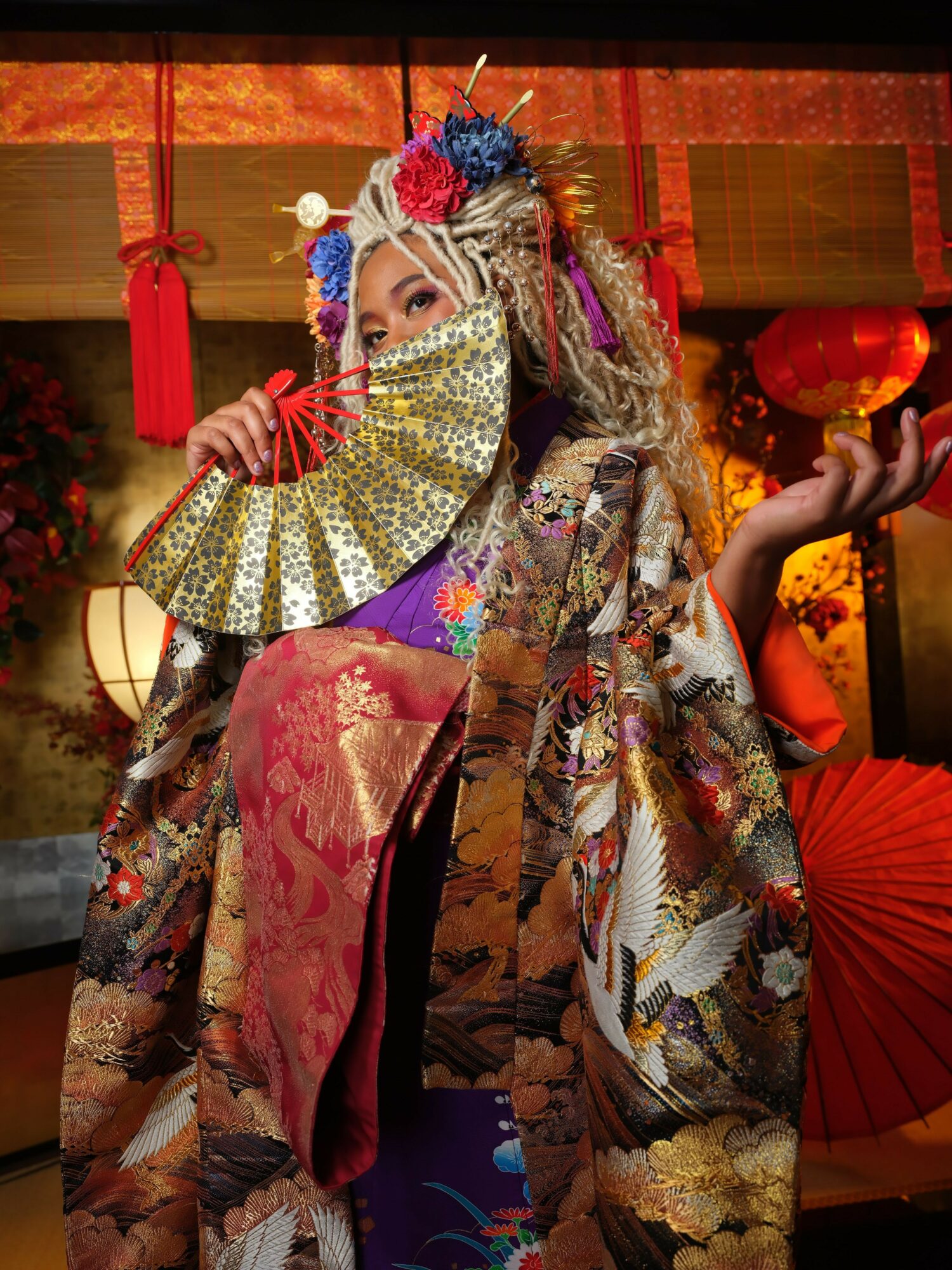 Person in traditional attire holding a fan, lying on a decorated surface with vibrant background elements.