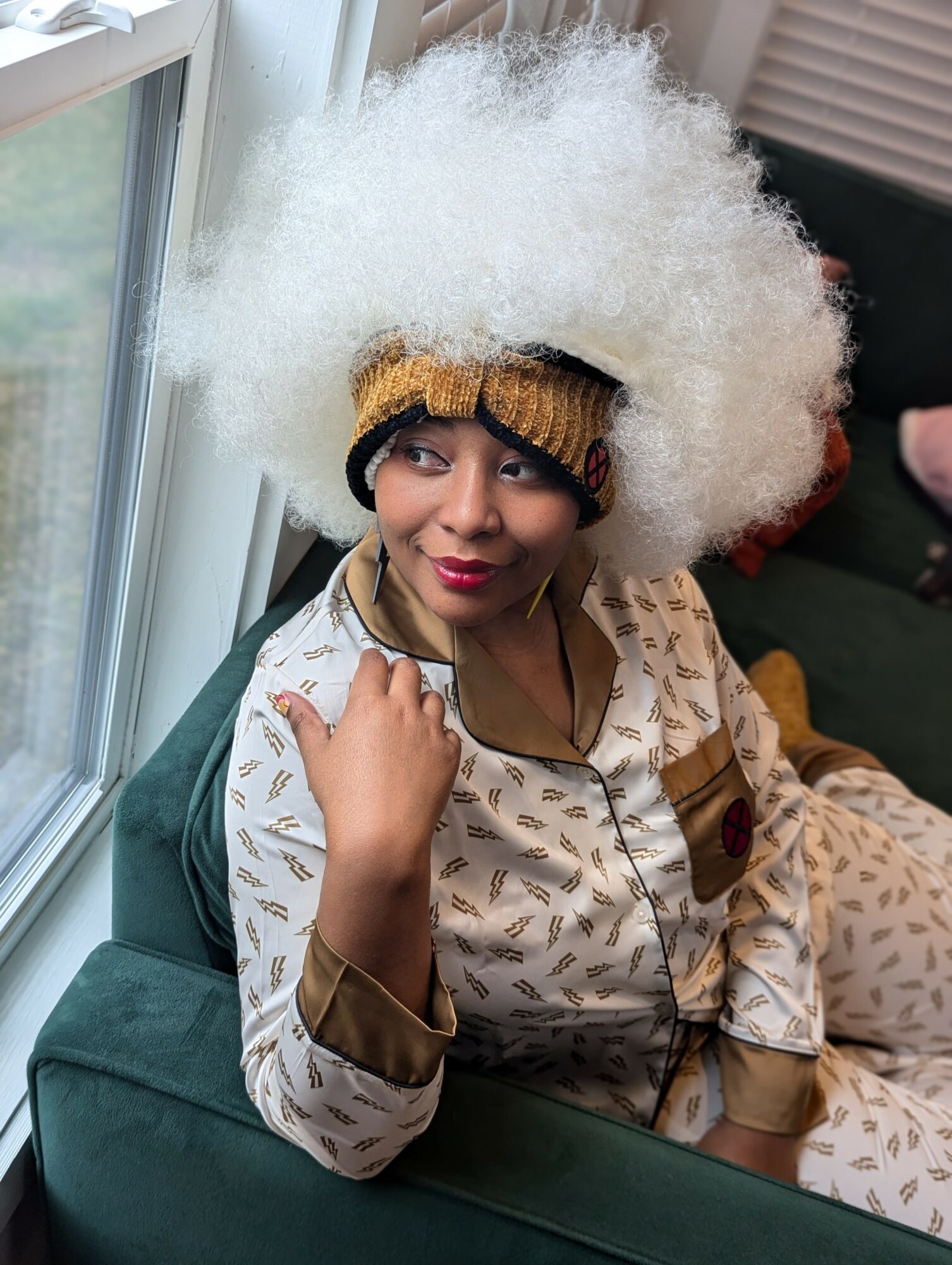 Woman with large white afro hairstyle sitting on a green couch, wearing patterned pajamas and a gold headband, near a window.