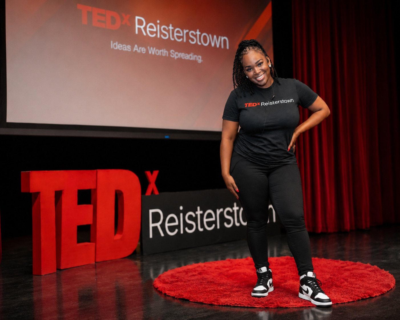 Woman standing on a red circular rug on stage, smiling, with TEDx Reisterstown sign and screen behind her.