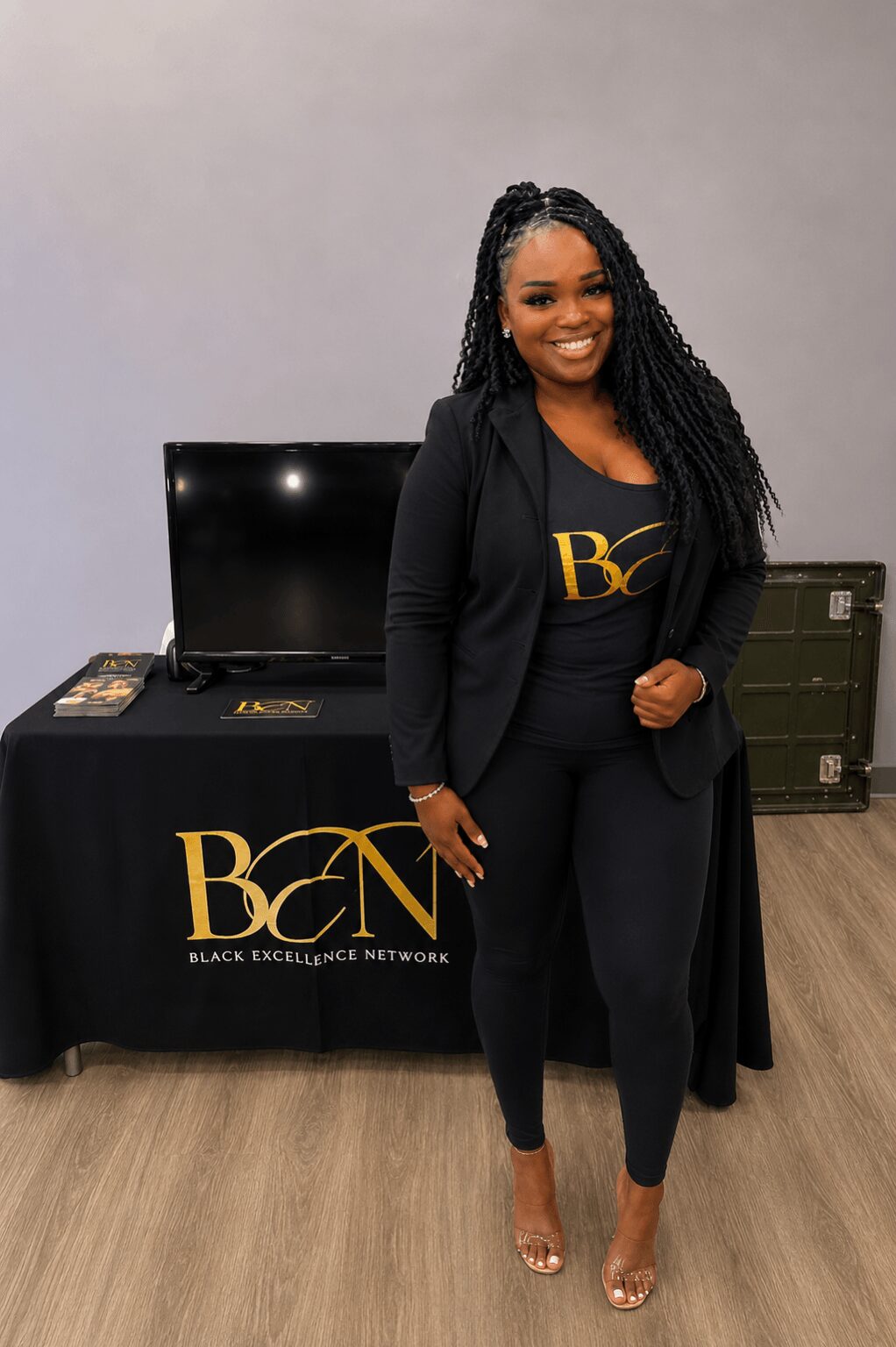 Woman with long braided hair smiling, standing in front of a table with a monitor and promotional materials, wearing a black blazer and outfit.