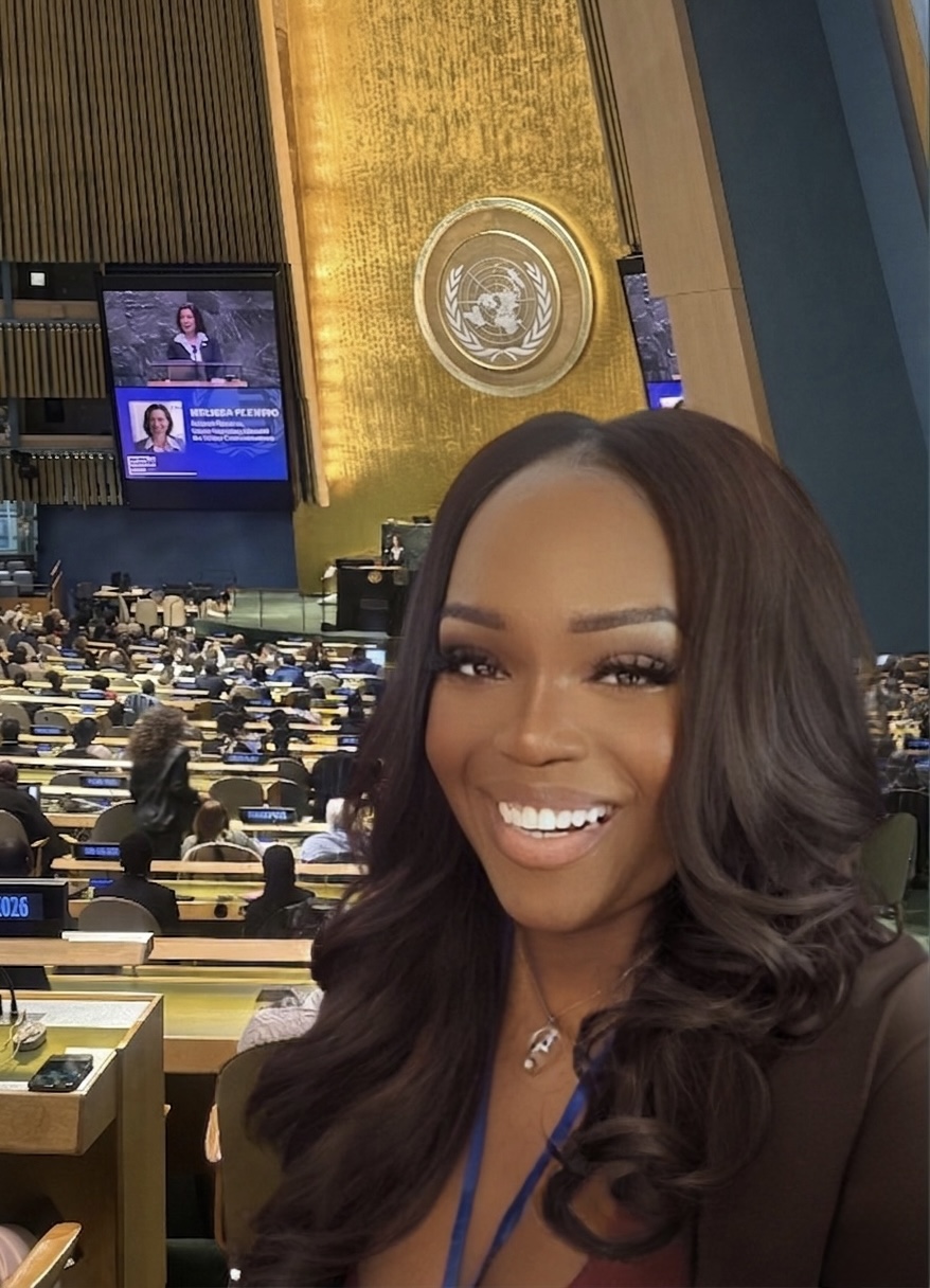 Smiling woman with long dark hair in a conference room with United Nations emblem on wall.