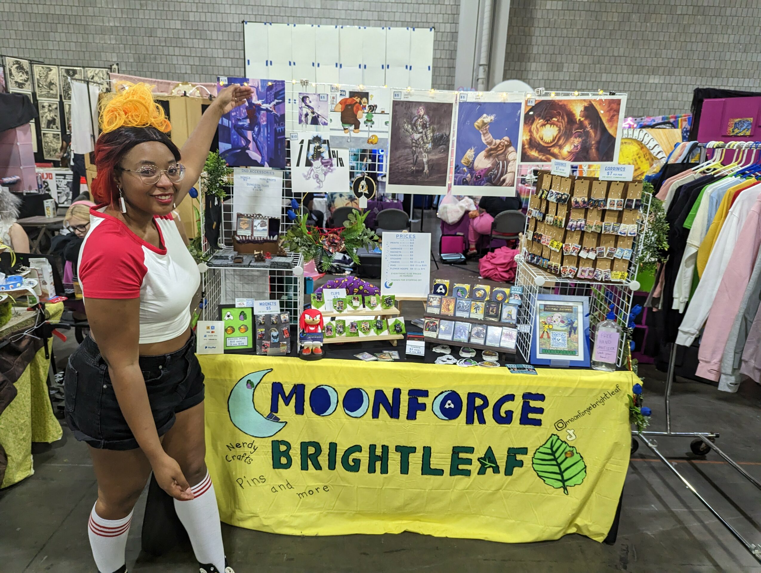 Person standing next to a booth with a yellow banner reading 'MOONFORCE BRIGHTLEAF,' displaying artwork and products at an indoor event.