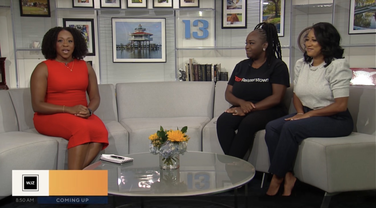 Three women sit on a white sofa in a TV studio, with a glass table and framed pictures behind them.
