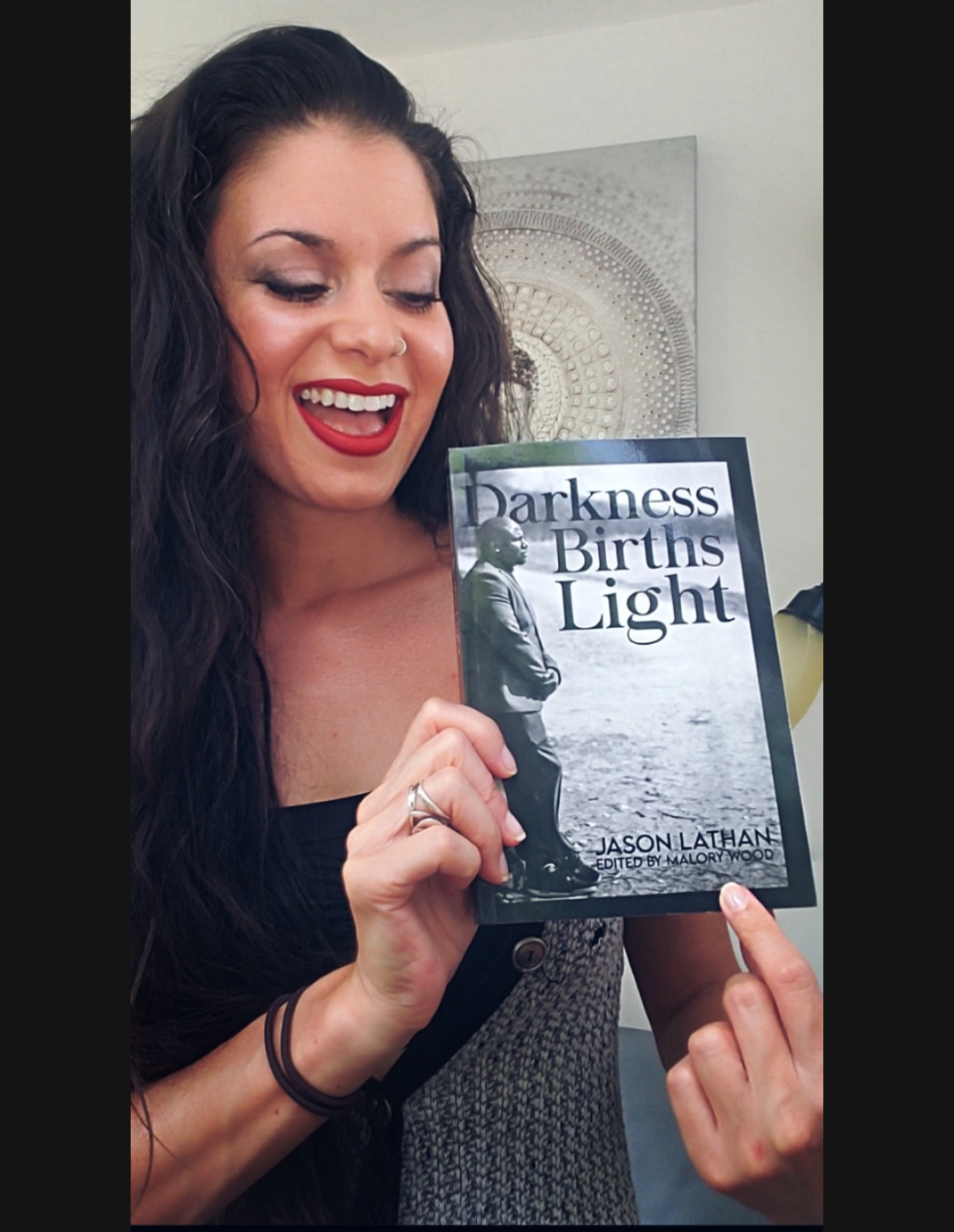 Woman with dark hair smiling and holding a book titled 'Darkness Births Light'.