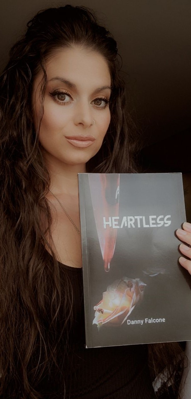 Young woman with long wavy hair holding a book titled 'Heartless' by Danny Falcone, looking at the camera.