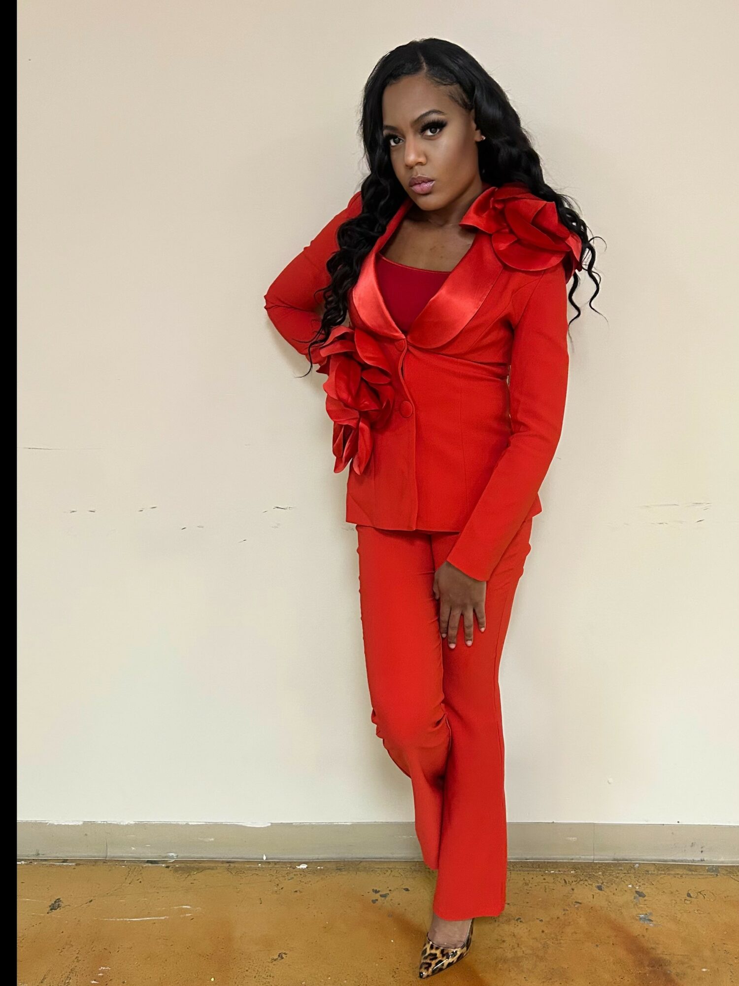 Woman in red suit with ruffled shoulder detail, standing against a plain wall, looking at the camera.