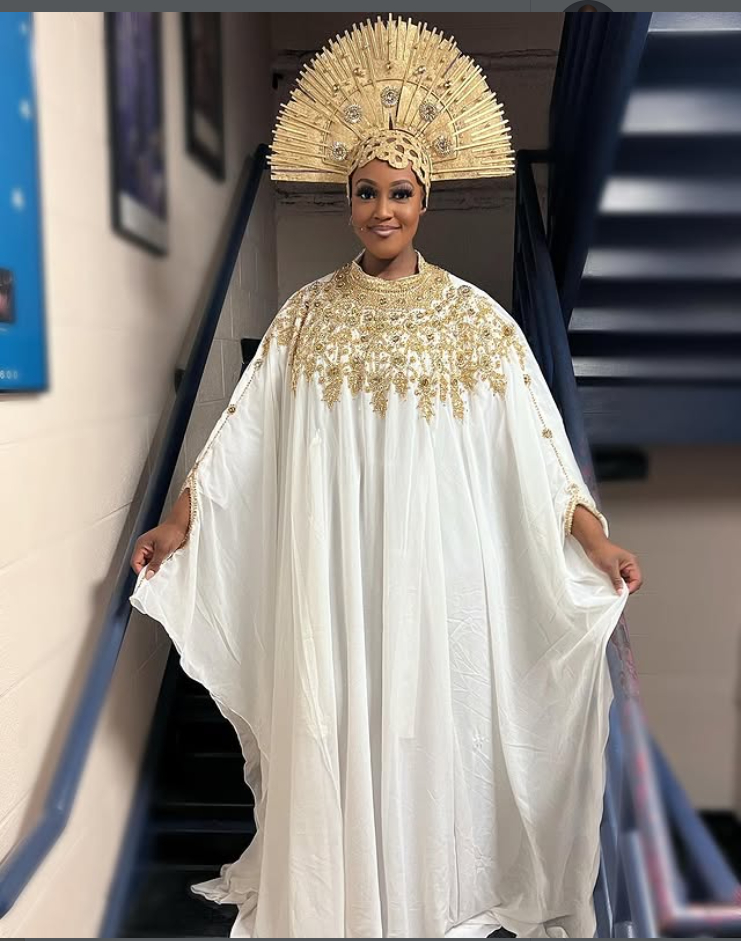 Person in elaborate white and gold costume with large gold headpiece, standing on stairs indoors.