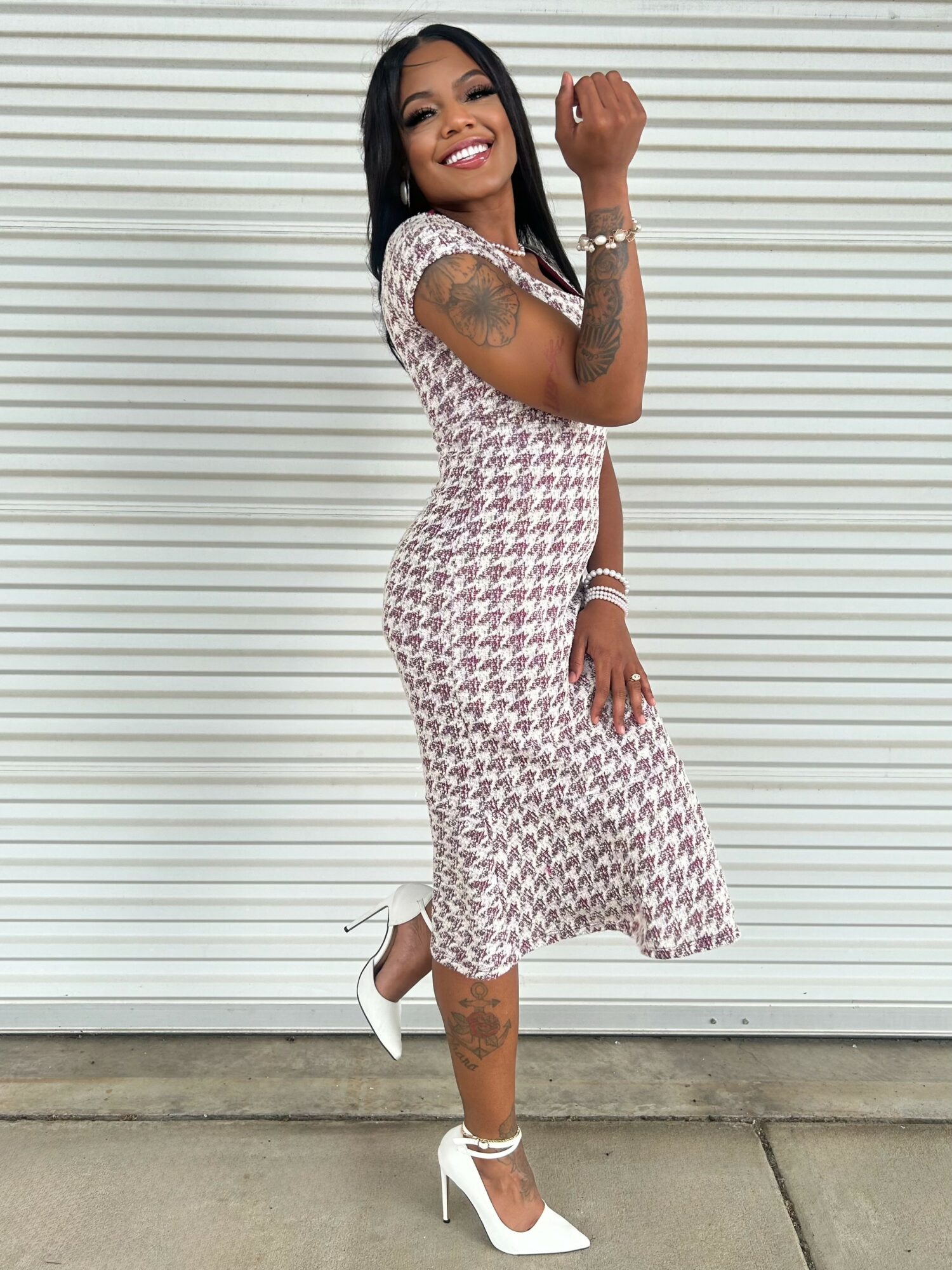 Smiling woman in patterned dress and white heels standing against a striped background, showing a fist gesture.