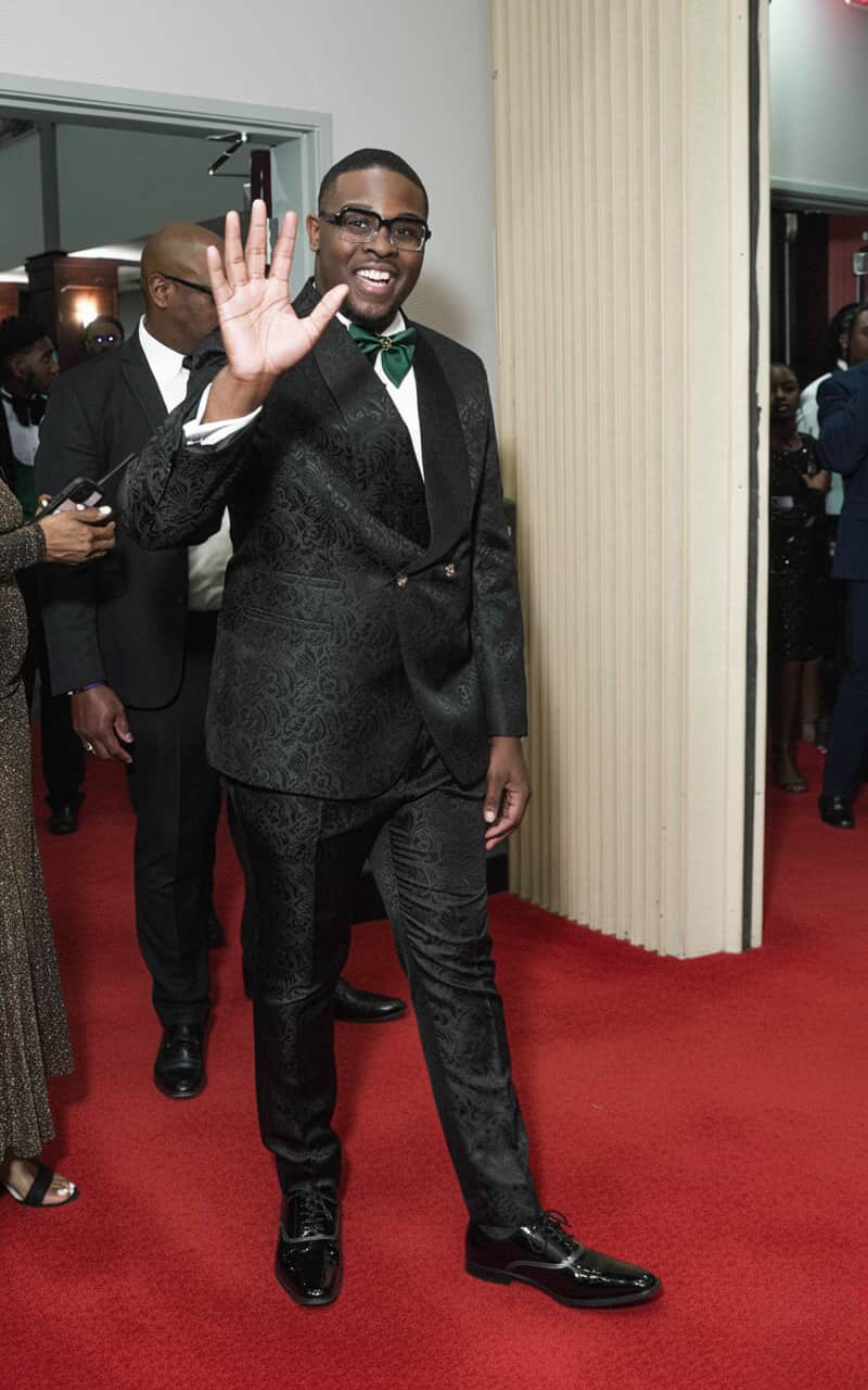 Man in a tuxedo waving and smiling on a red carpet, with people in the background.