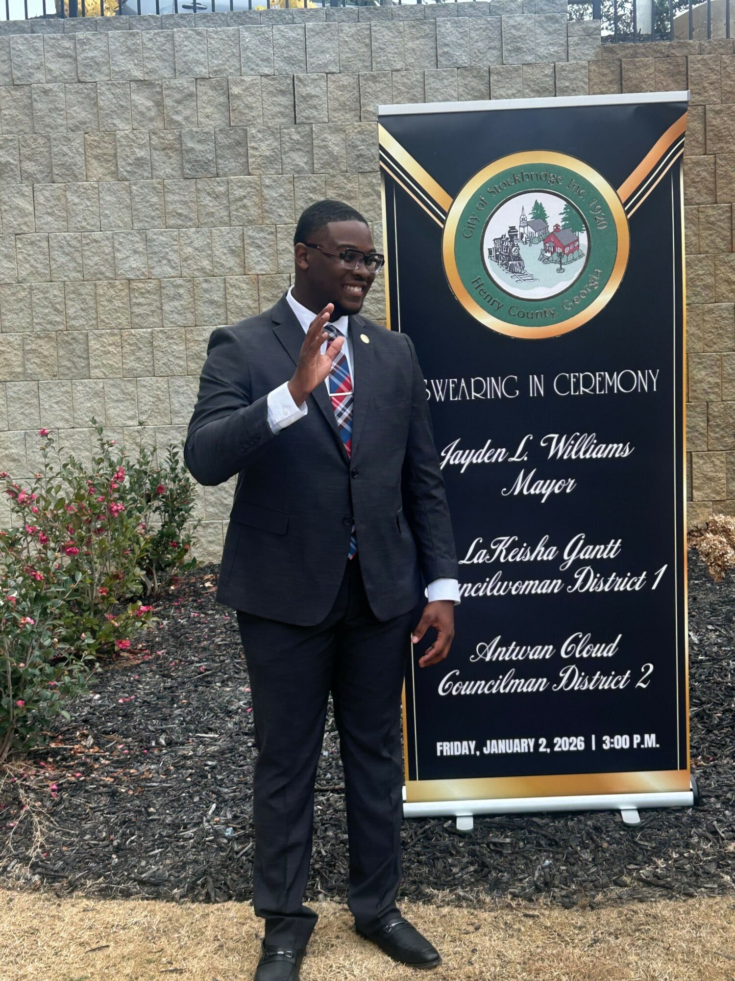 Man in a suit talking on a phone, standing outdoors near a sign with text and a logo.