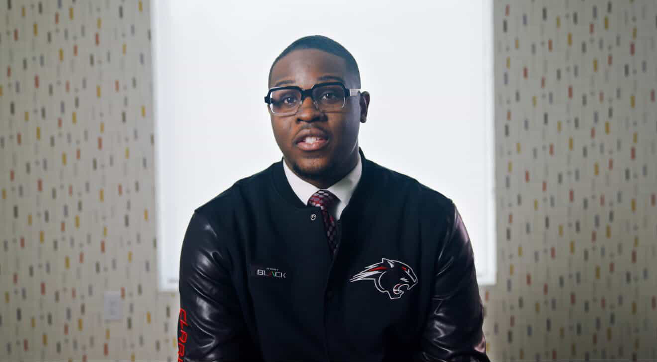 Young man wearing glasses and a black jacket with a Baltimore Ravens logo, standing between patterned walls.