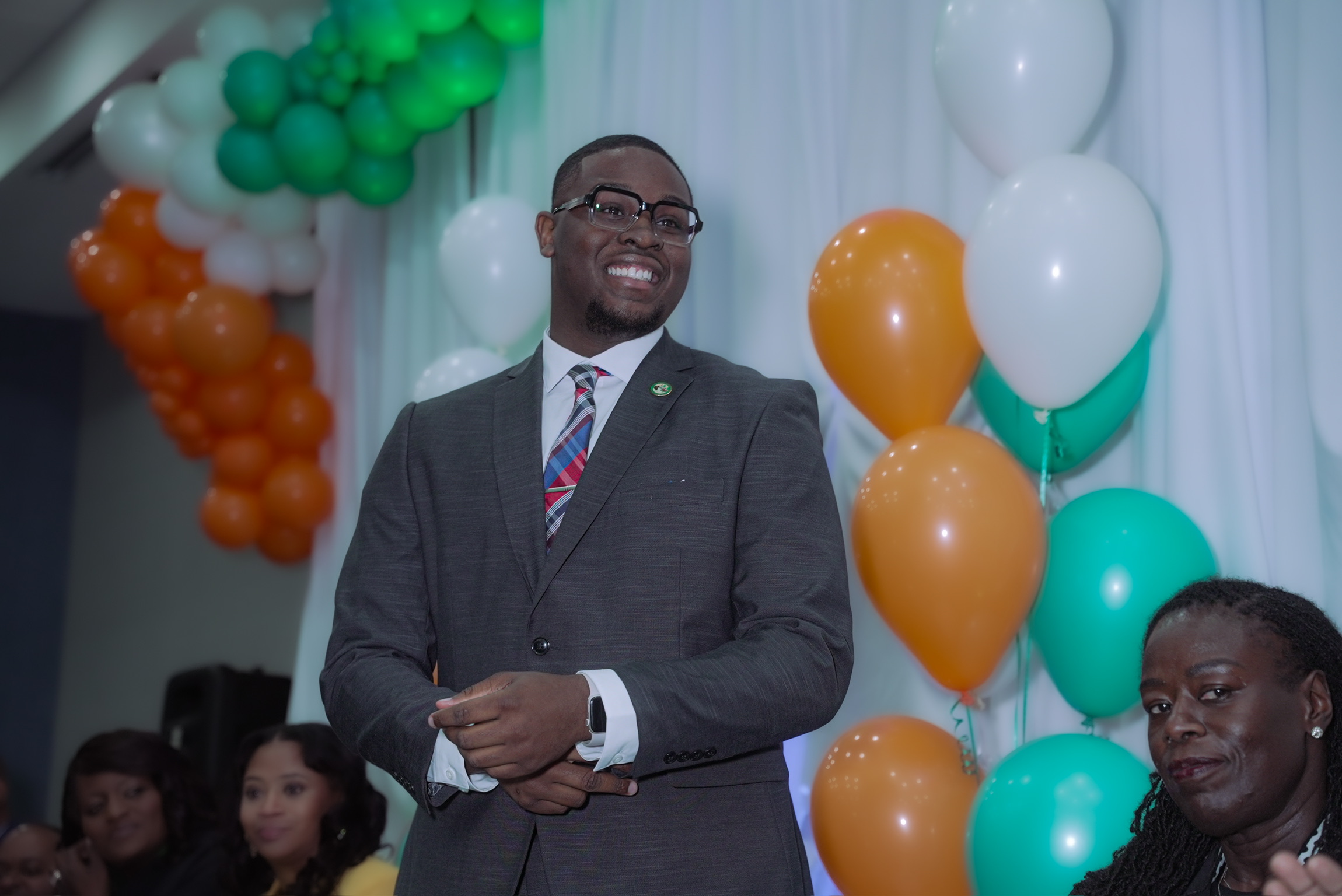 Man in suit smiling at celebration with colorful balloons and white drapes in background.