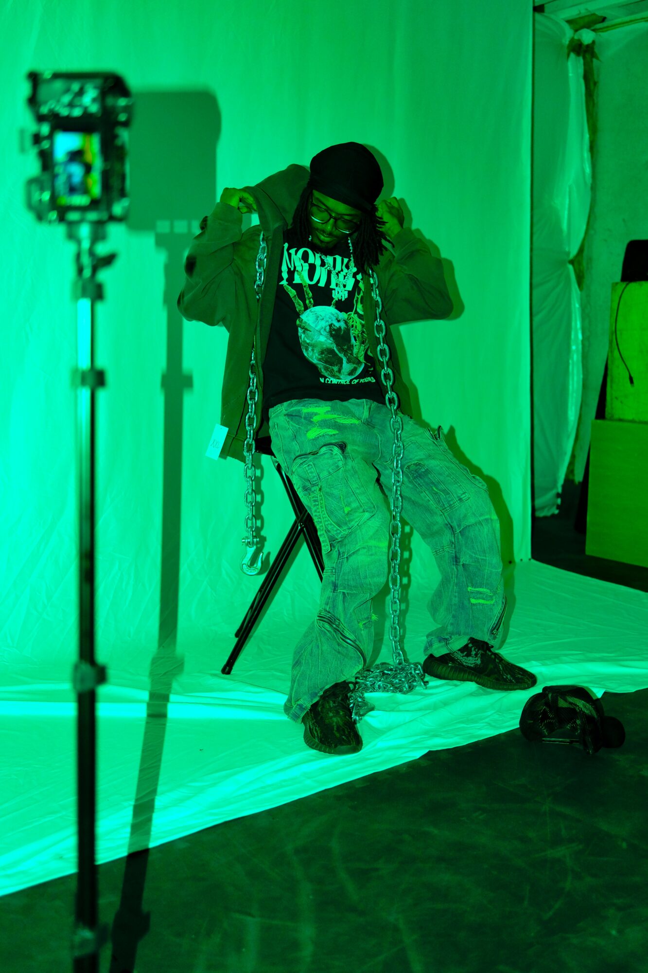 Person sitting on a chair with a green background, wearing a jacket, graphic t-shirt, and ripped jeans, adjusting their hood.