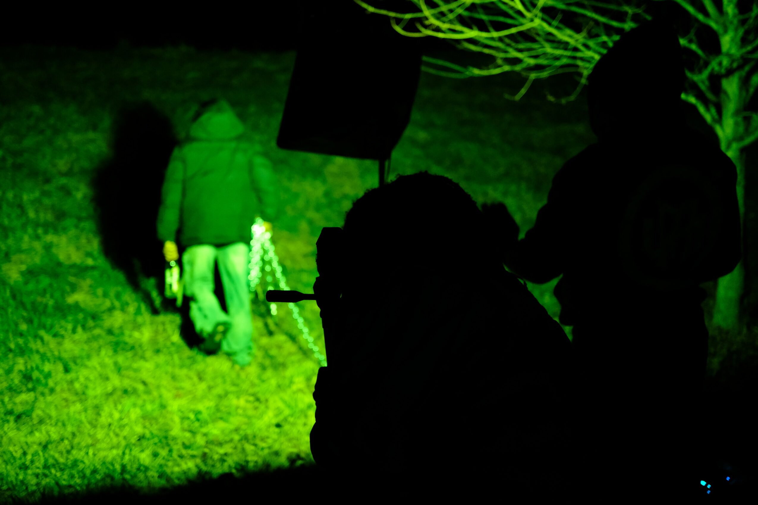 Two people with backpacks and a person with a hooded jacket are outdoors at night, illuminated by green light, with trees in the background.