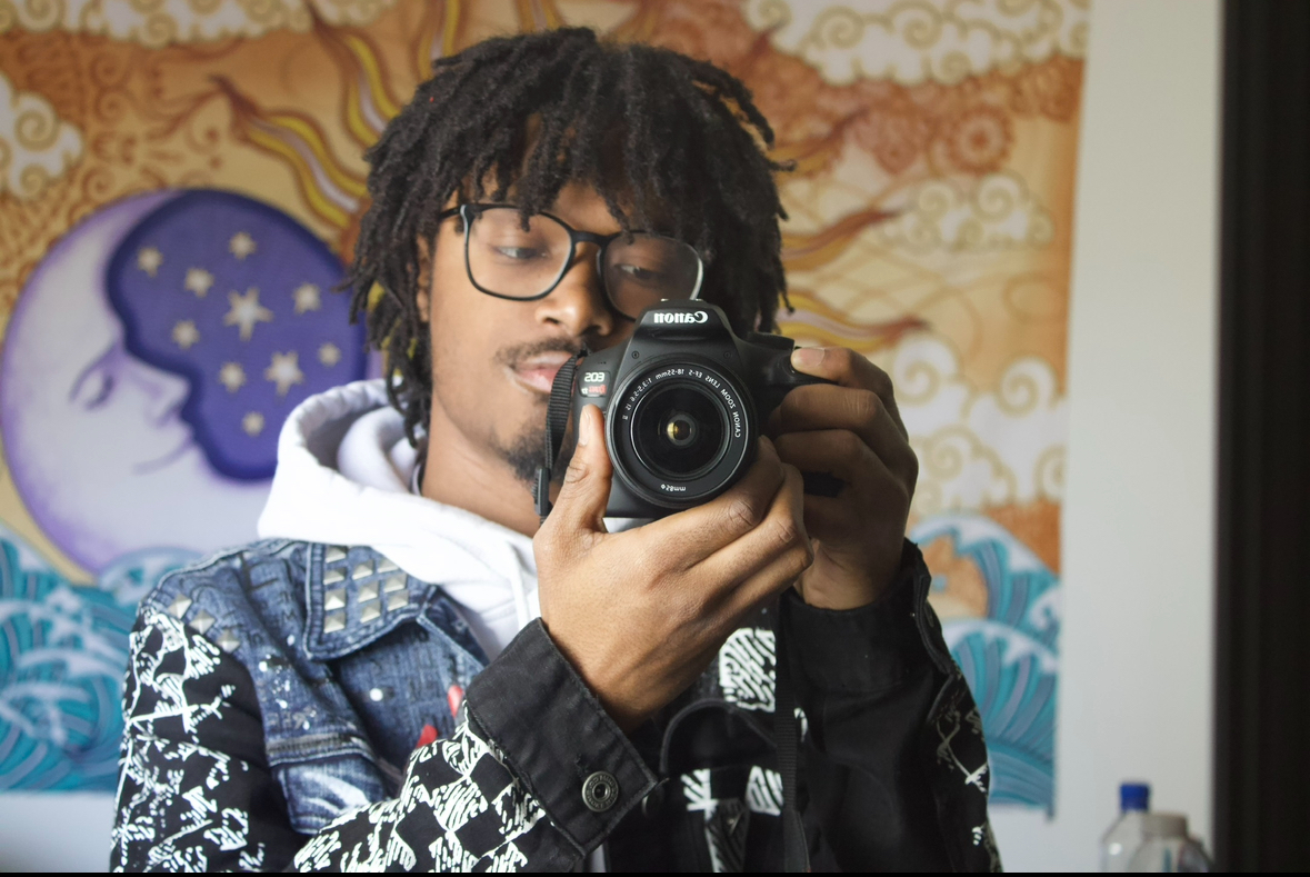 Person with glasses and dreadlocks taking a selfie in front of a colorful wall, holding a camera, wearing a patterned jacket.