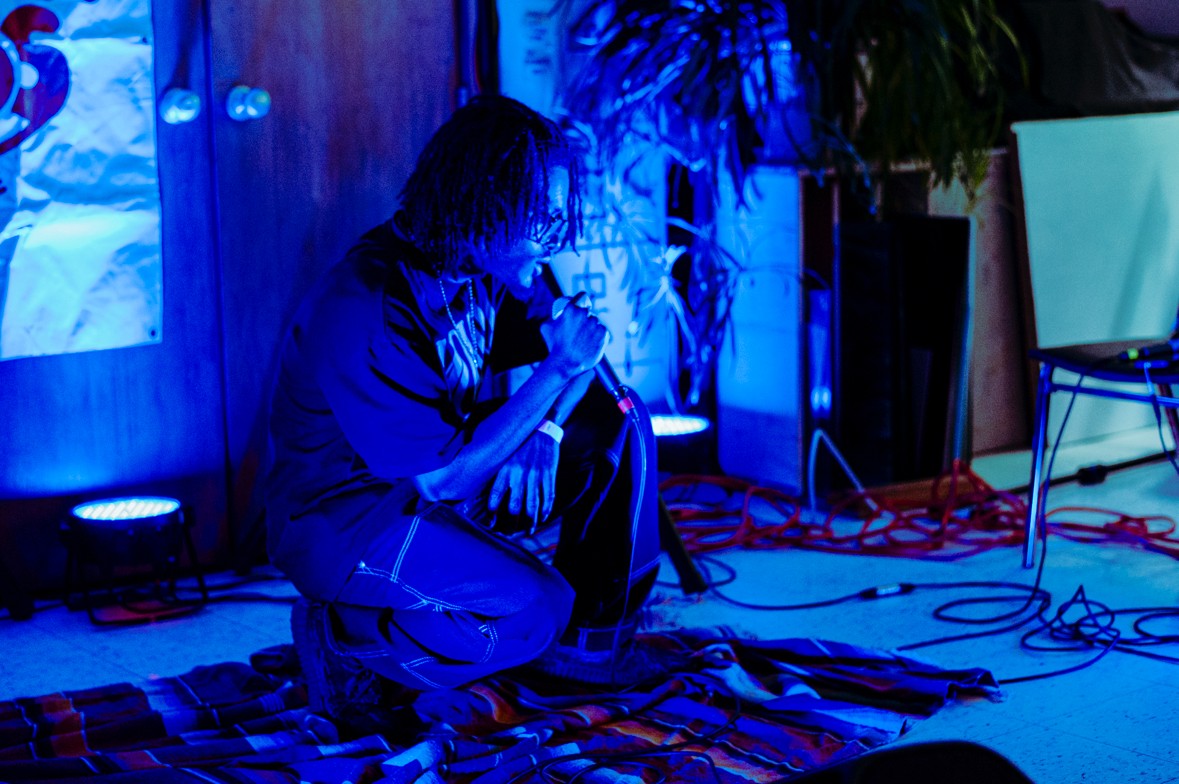Person sitting on the floor holding a microphone, surrounded by cables and equipment, with blue lighting and plants nearby.