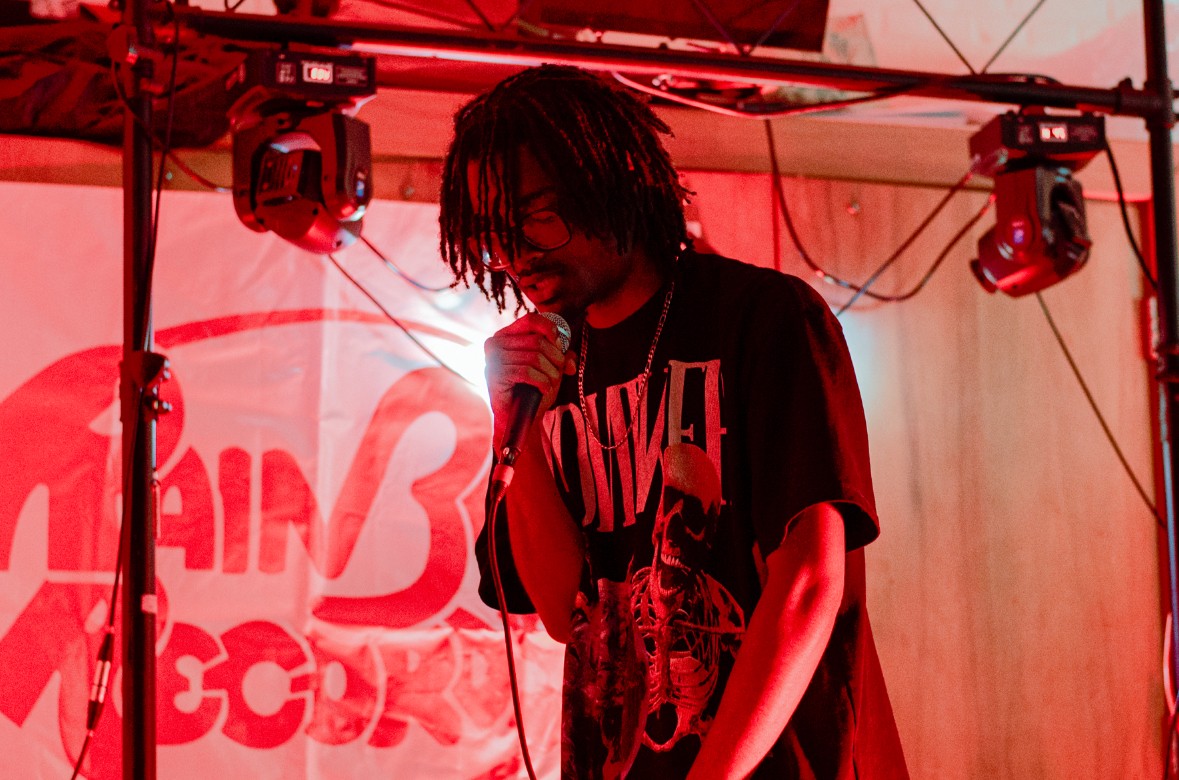 Person with dark hair singing into a microphone on stage with red lighting and equipment overhead.