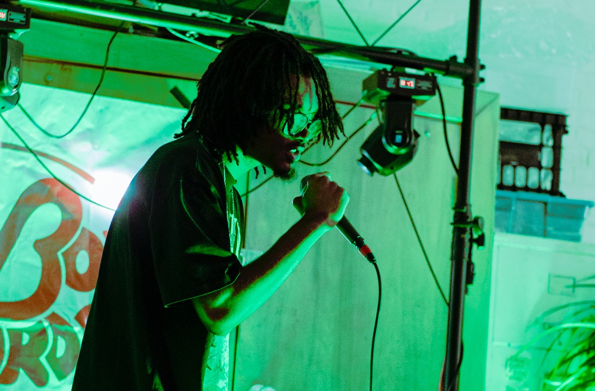 Person with dreadlocks singing into a microphone on stage, green lighting, equipment visible in background.