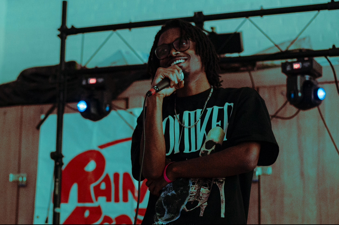 Person with glasses and dreadlocks holding a microphone on stage, with a colorful background and stage lights.