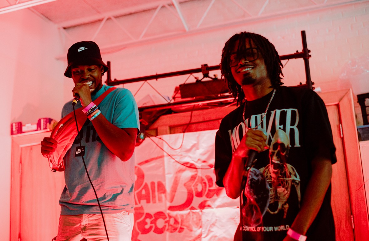 Two people holding microphones on stage with a red background and equipment behind them.